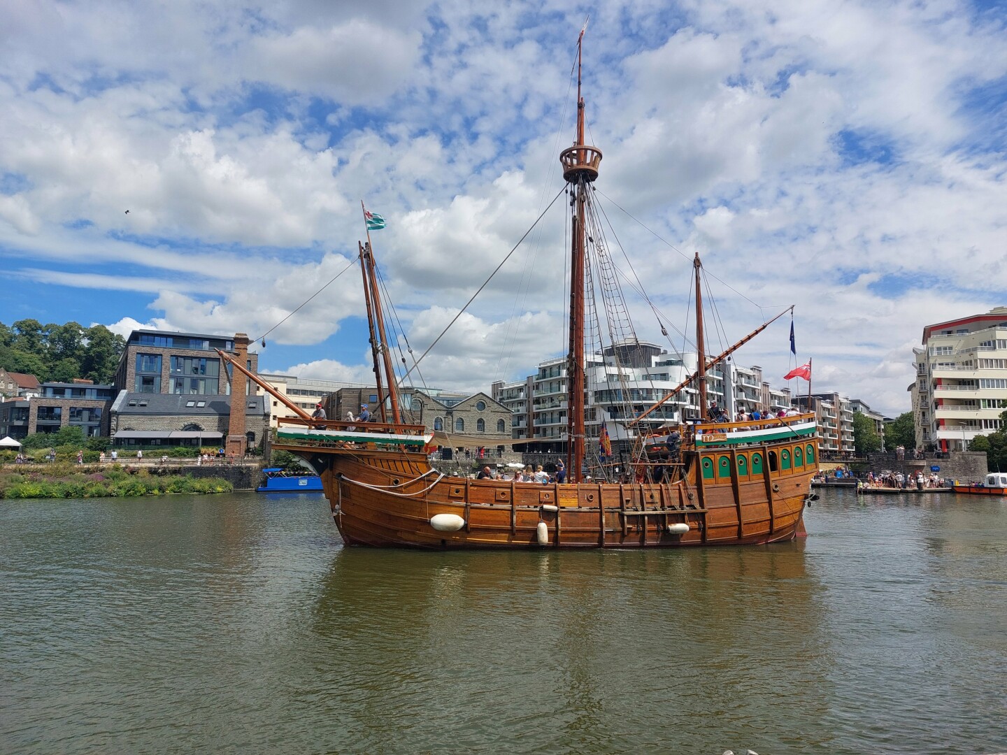 A large brown wooden mast boat sailing in front of office buildings. Set against a cloudy blue sky.