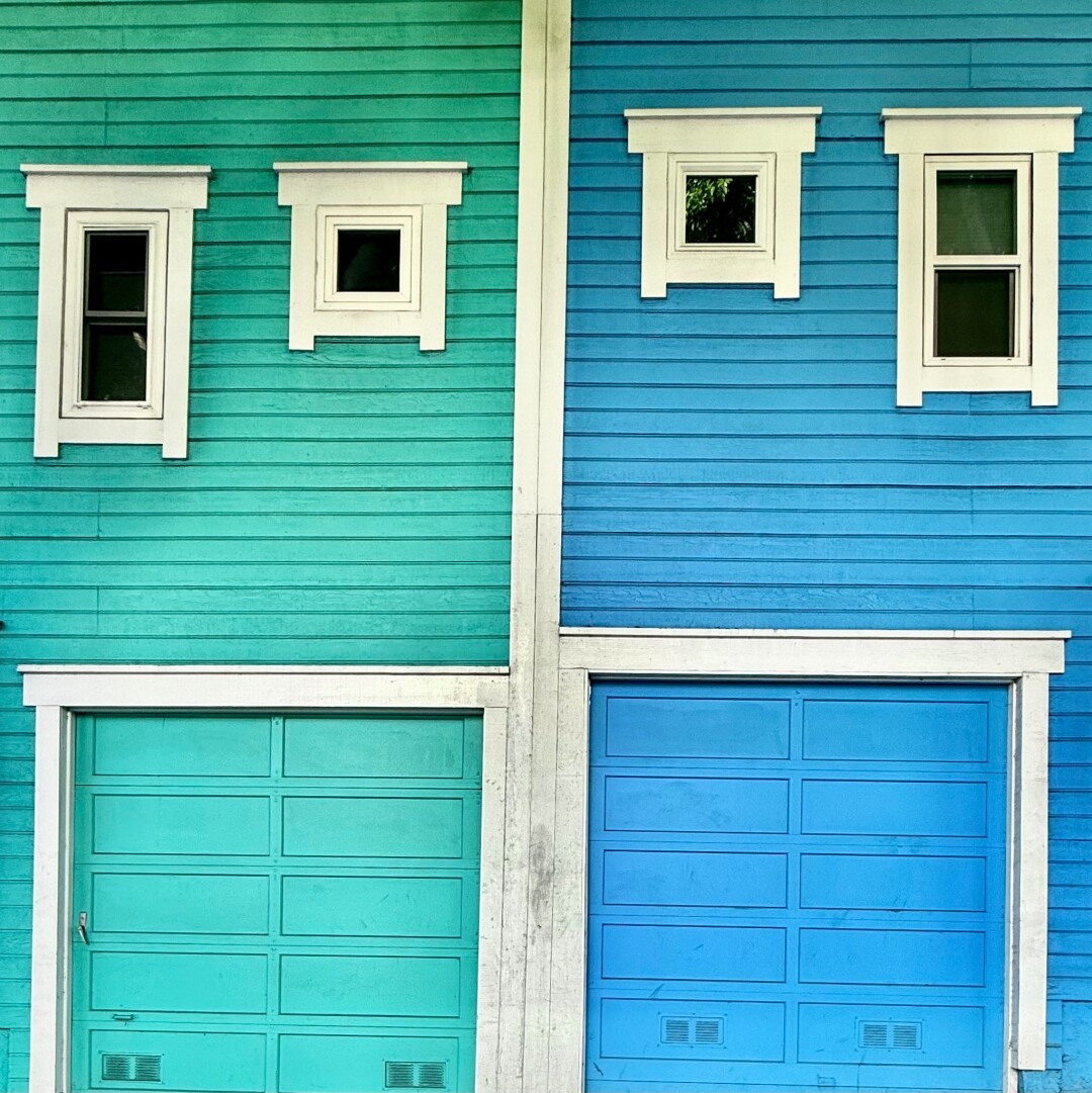 Side-by-side dwellings. Both have one small window and one larger window over a garage door, but they're mirrored kind of making them look like the faces in that dumb lel emoji. The left side is painted kind of a darker mint green and the right is more of a cornflower blue. Both have white trim.