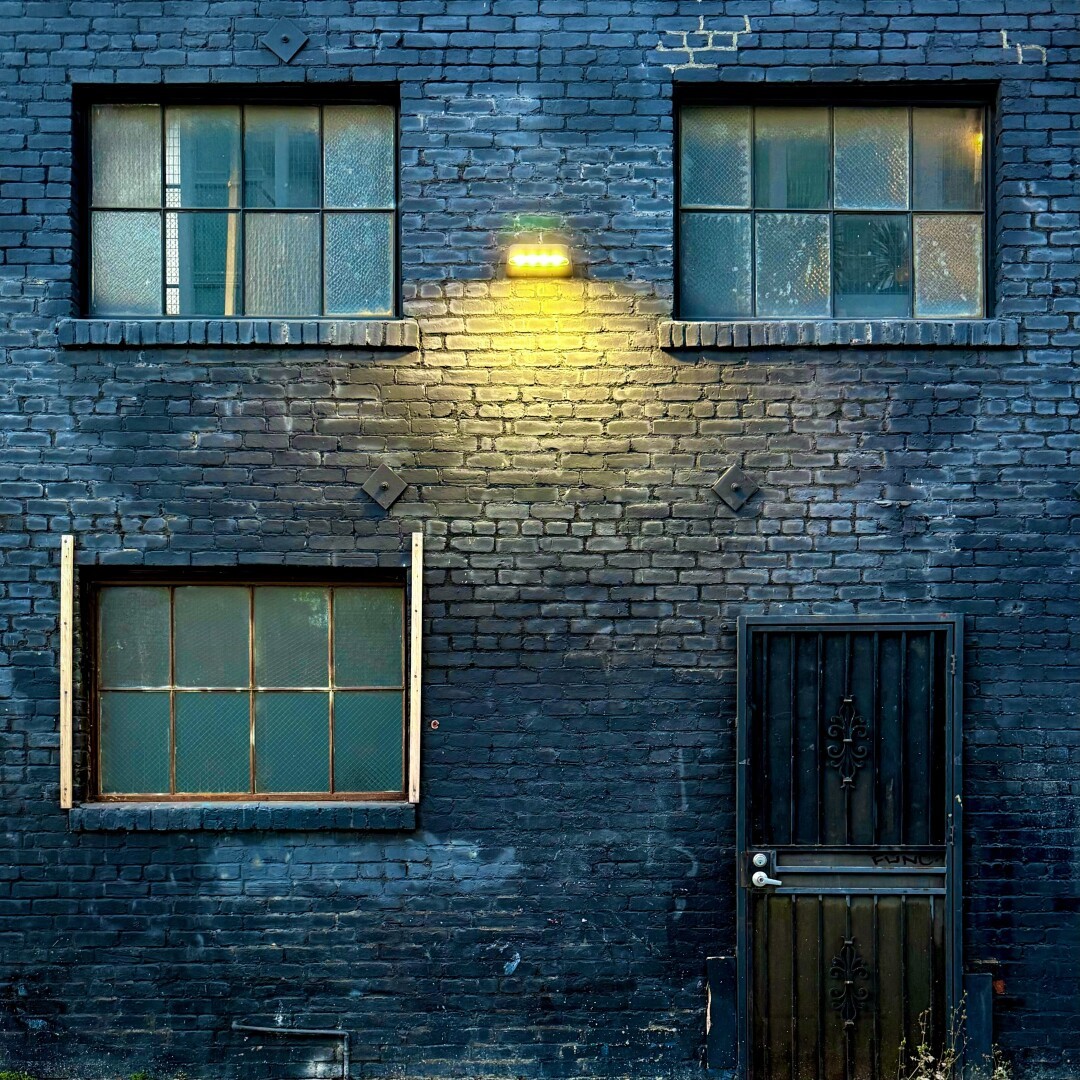 Exterior wall of a dark brick building with factory windows and a gated door. There is a single yellow light illuminating the wall, making everything else have a blue-green tint.