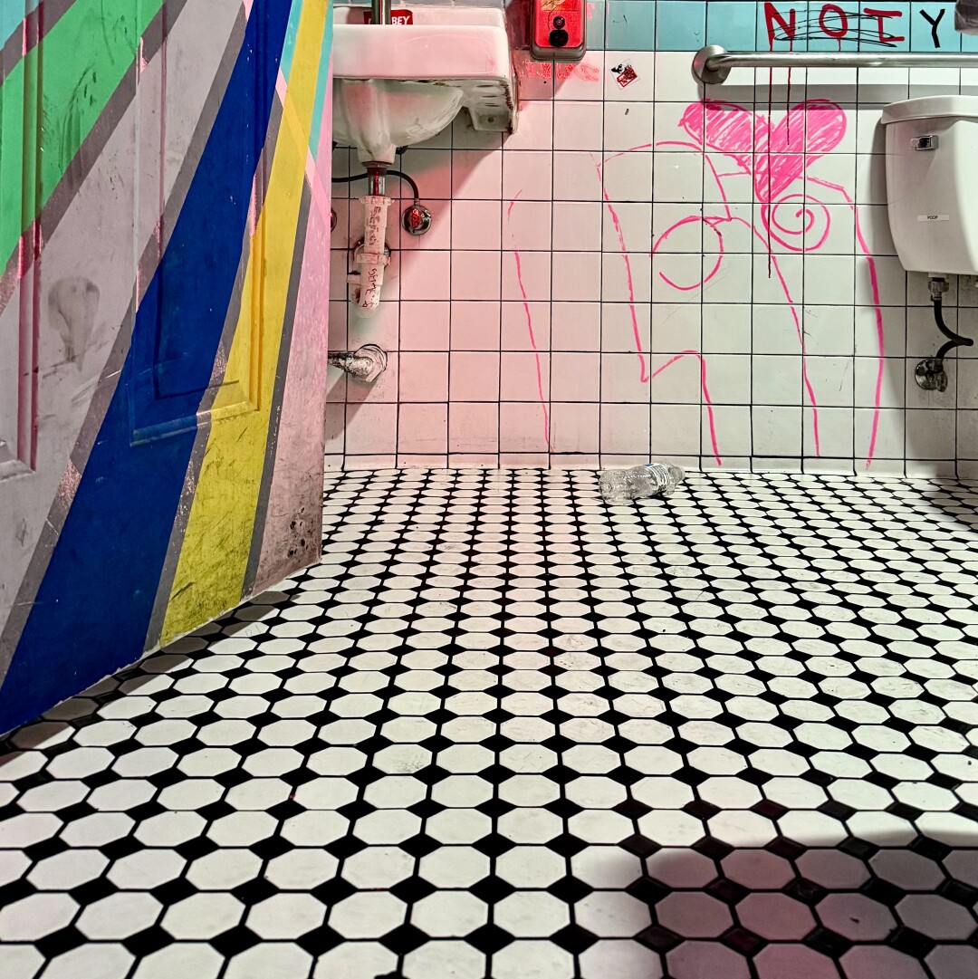 Low angled shot of a bathroom floor in a bar. There is white and black tile and a door painted in pink, yellow, blue, and green lines radiating out from around the door handle