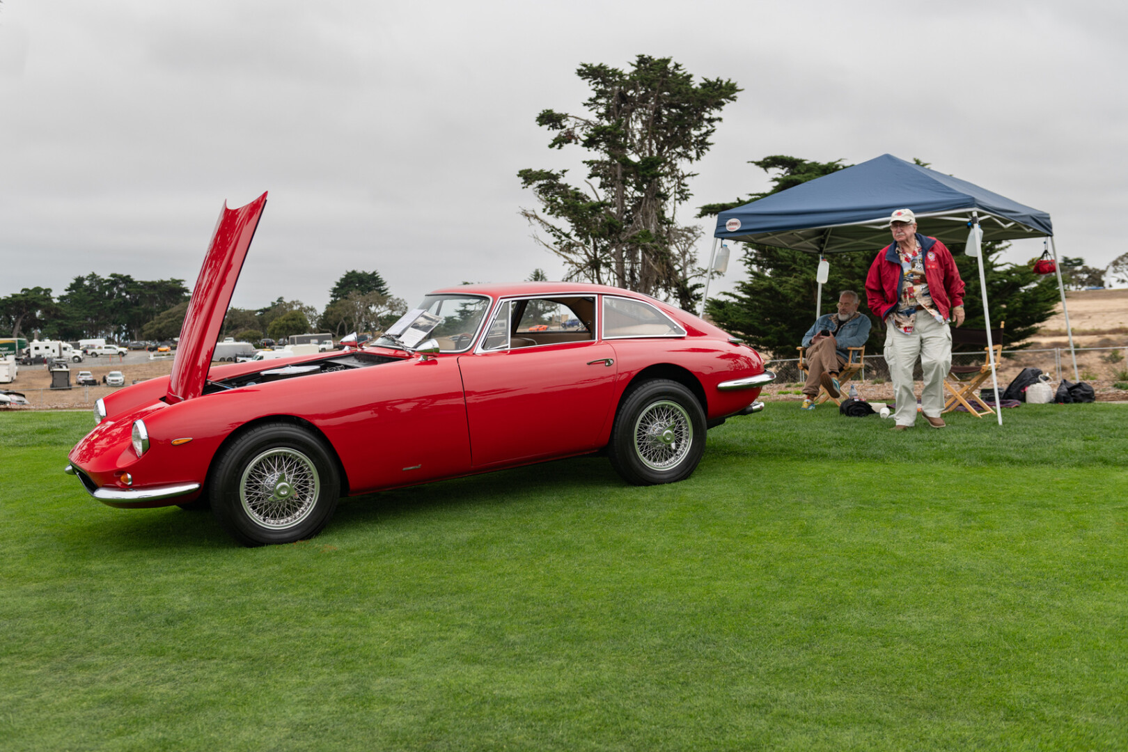 A red long-hooded vintage coupe, parked on a golf green by a temporary tent housing a couple of people.