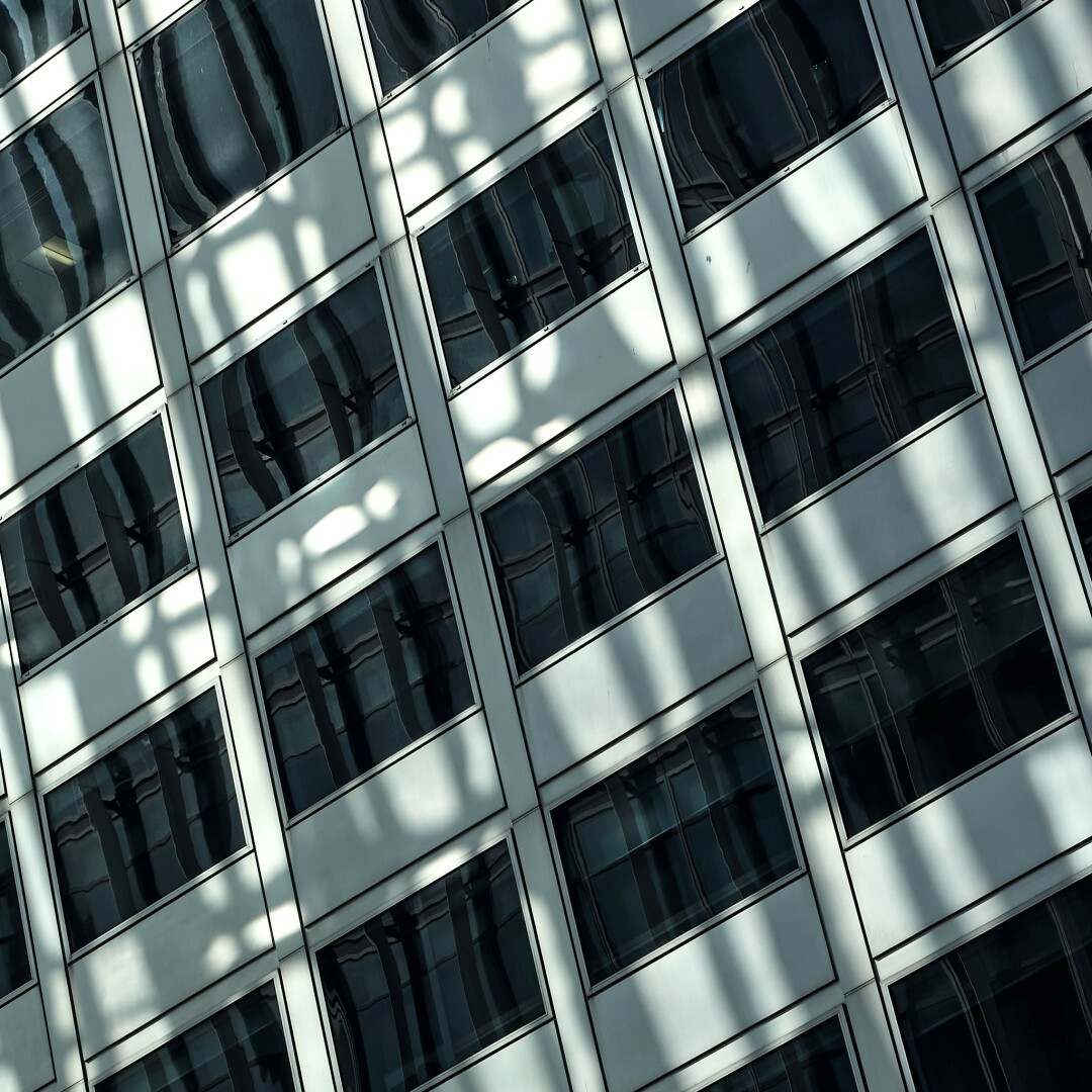 Tilted photo of dark wide rectangular windows in a white plastic-looking wall which wavily reflect a building across the way, narrow windows behind vertical slats. Brighter light from this other building's is cast onto the first, this reflection overlapping and in some cases meshing with the ones in the glass.