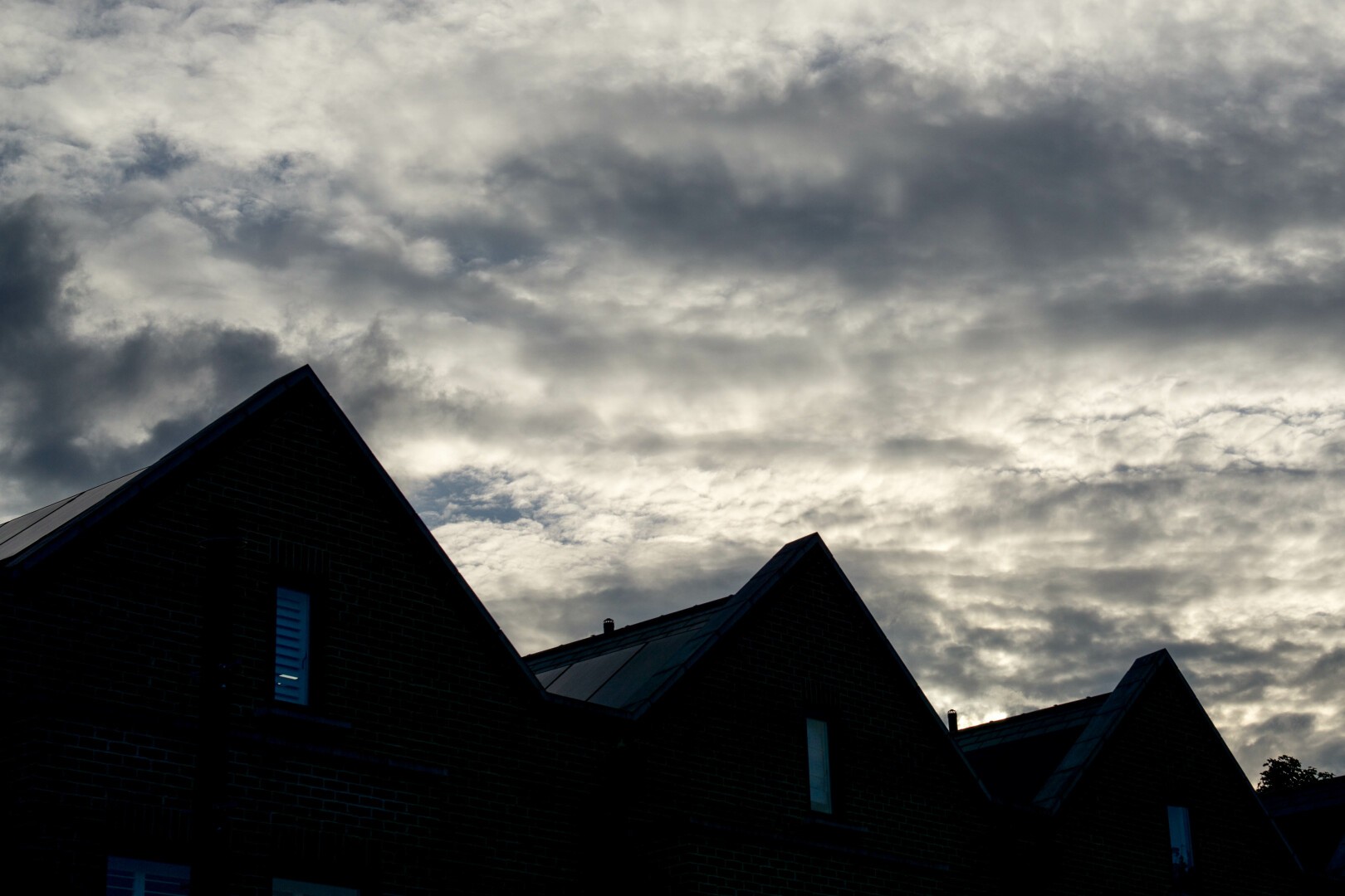 Photo of three gable ends silhouetted against a bright sky covered with rumpled grey-white cloud. Just about visible in each of the dark triangles is a slim white window. The small slices of roof visible above them shine dully with reflected light.