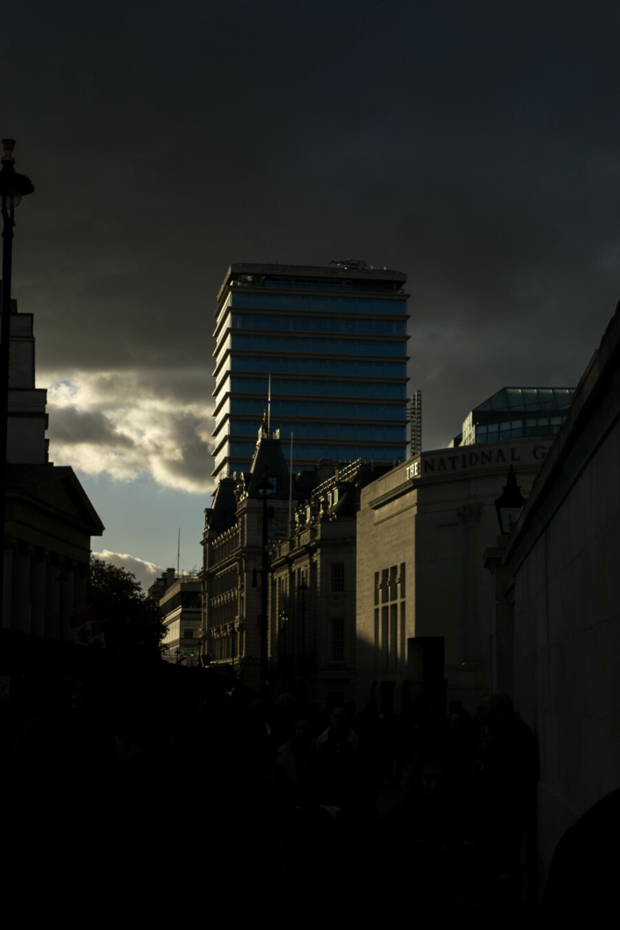 Photo looking along a city street, angled slightly upwards towards dark clouds, low sunlight coming in from the left somewhere. The deep shadows at the bottom of the image encompass everything on the left of the road and cut a jagged line across the buildings on its right, their lower floors and the road in front of them invisible in the darkness. In the middle distance the buildings are several stories tall and made of light brick and stone, the nearest has the word 'THE' on its closest corner. Behind them is a moderately tall glass and concrete office block, left face lit up like someone whose lover has just entered the room. Beyond that, the clouds break raggedly, revealing a patch of grey-blue sky. This was taken from the corner of Traflagar Square in London, looking west past the modern entrance of The National Gallery.