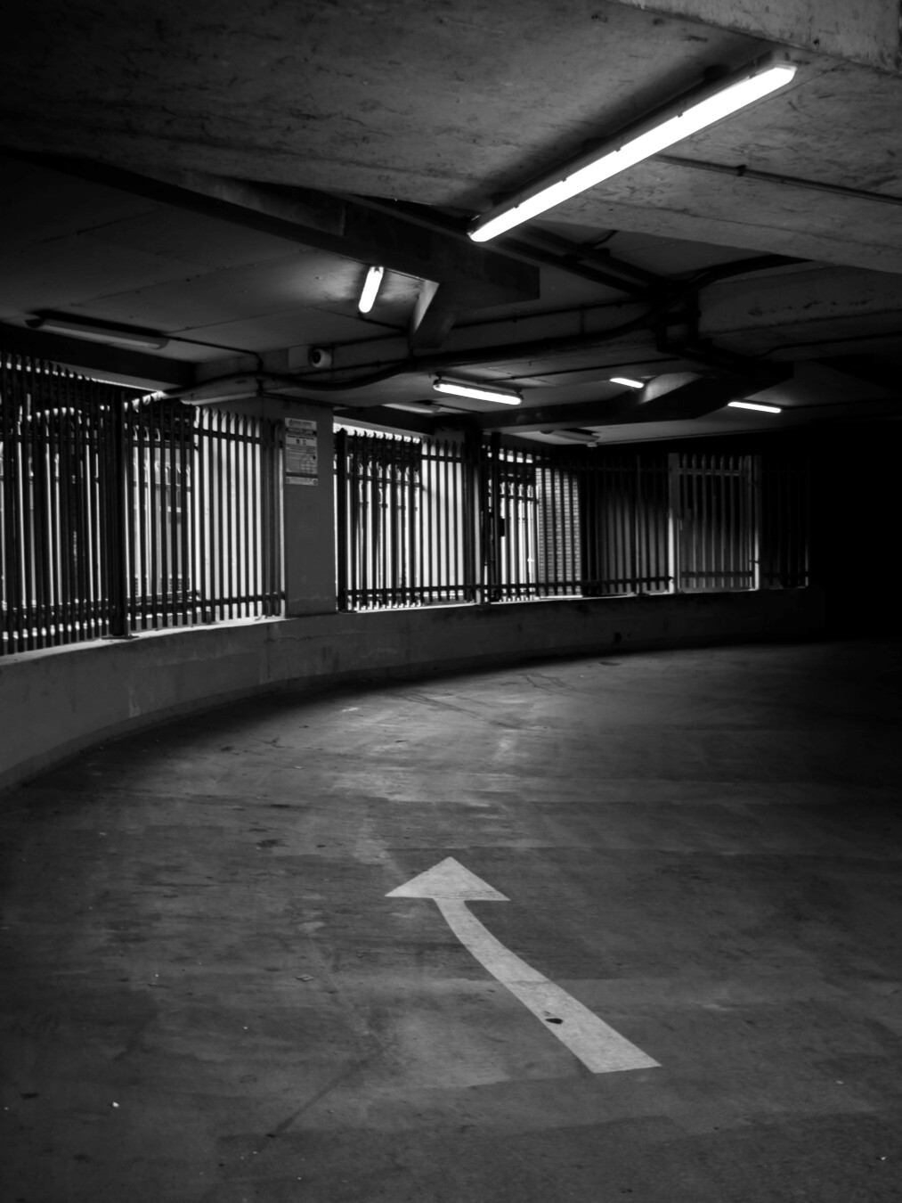 Monochrome photo of the inside of a car park entrance. A grey ramp curves up and to the right, a white arrow painted on it showing the direction to travel. The left side is bounded by tall dark railings in a concrete wall, a concrete ceiling above carries long straight lights that follow the ramp, shadows encroach from the right.