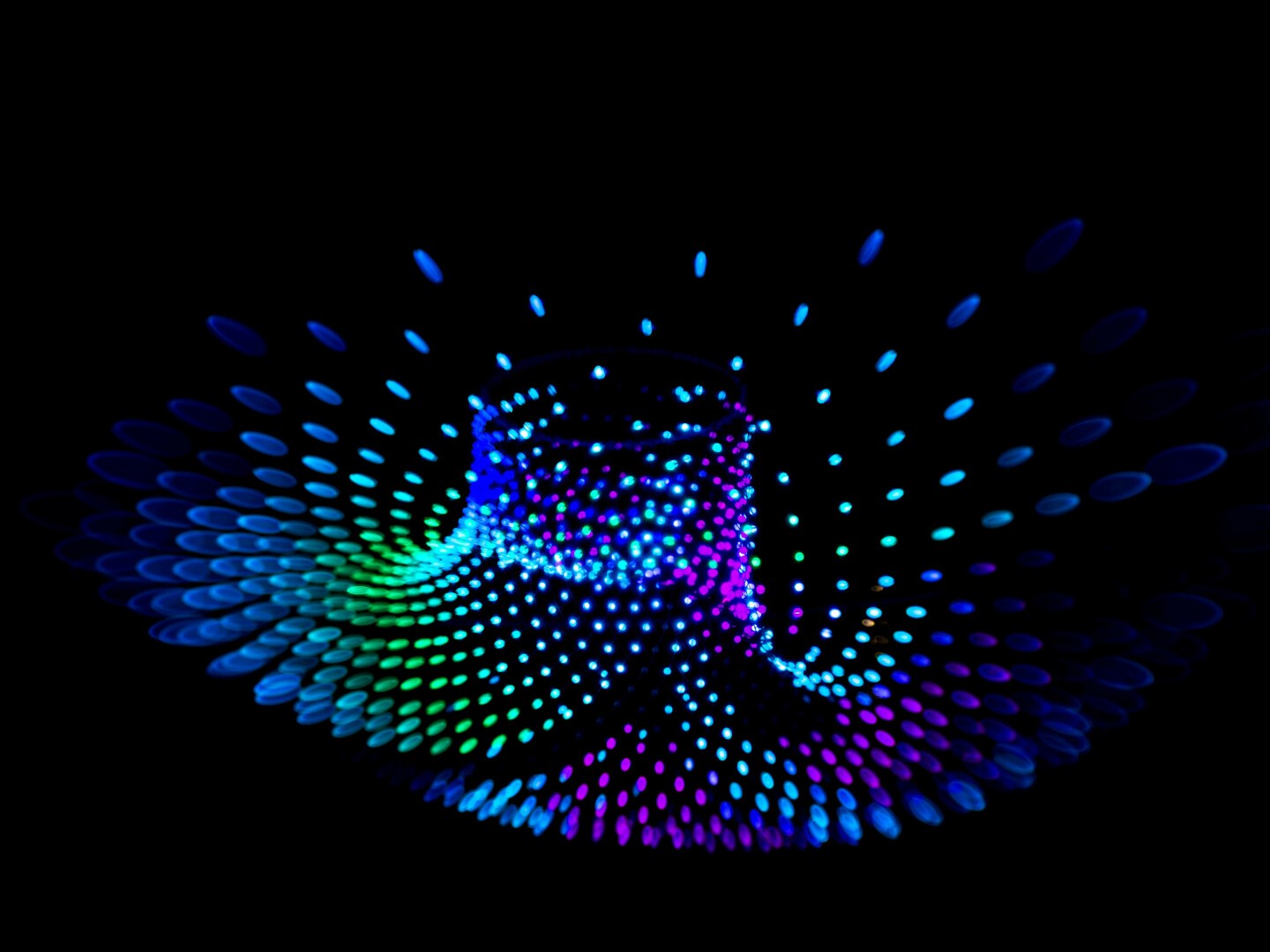 Photo of many lights, blue, green and sometimes violet, taken against a dark background, suspended in the air, blurred and stretched the further they are from the centre. In this image they appear to be in the shape of a skirt, caught mid-twirl, edges swung out and up.