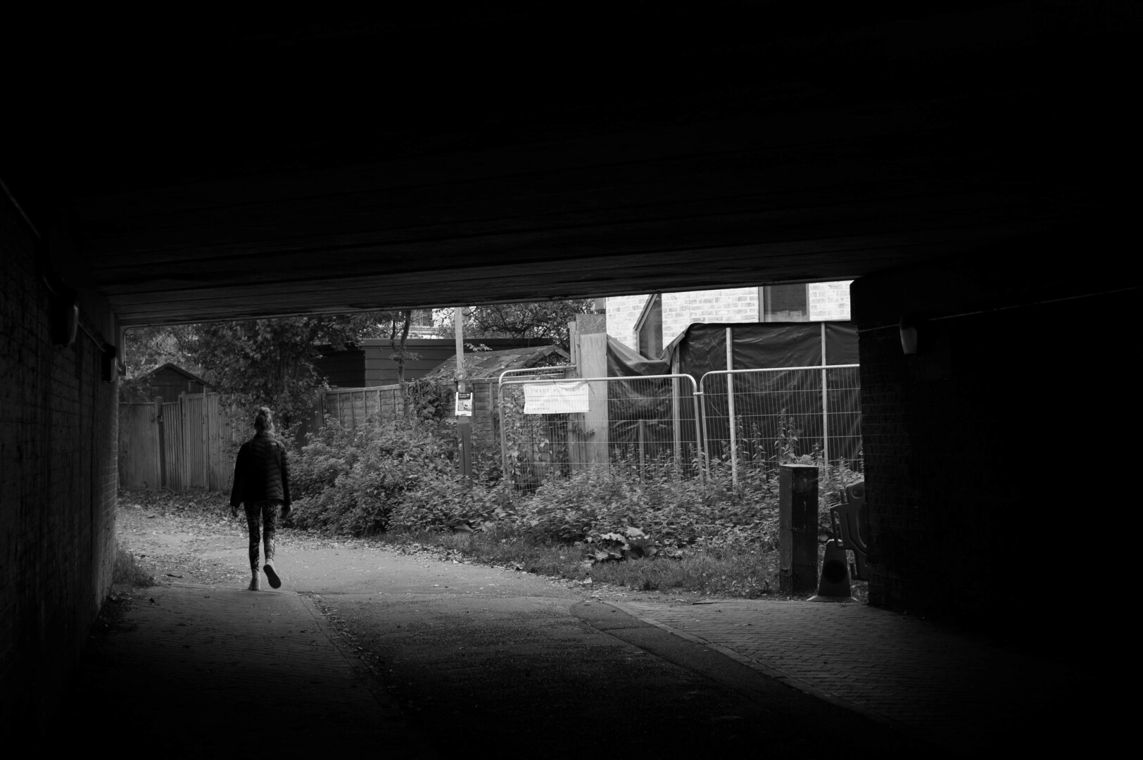 Monochrome photo of a lone figure walking out from under a rectangular dark enclosed space, taken from within that space. The path heads to the left, on the right is a slightly chaotic scene of wooden and temporary metal fencing, black plastic sheeting, an overgrown verge and glimpses of sheds and a newly-built building.