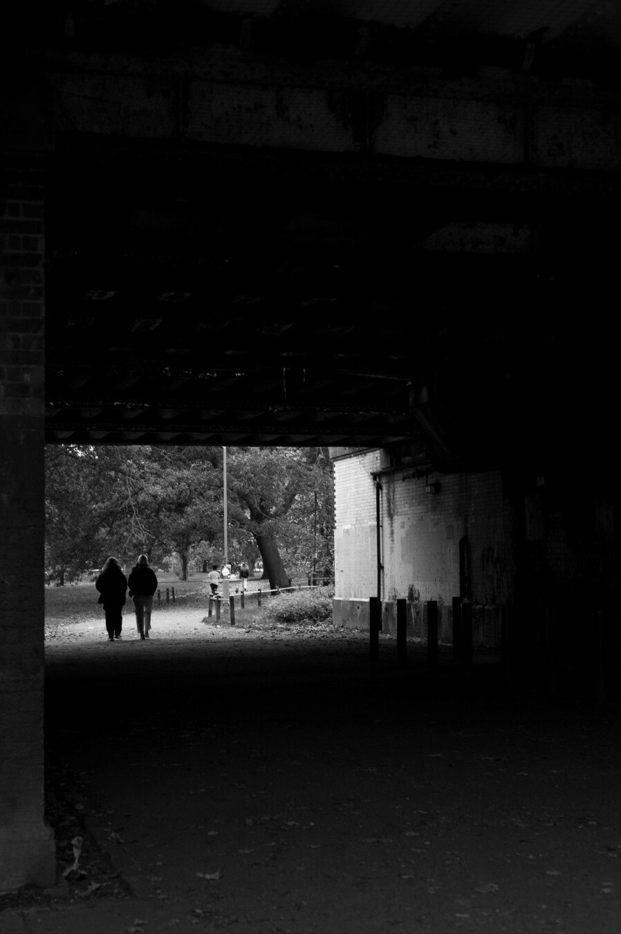 Black and white photo in which two people are on a path walking away from the camera are leaving a wide, rectangular, enclosed space and entering an area with several mature trees. Most of the photos is of the inside of the space, which is pitch black compared to the outside world, which is just a slim bar of light.