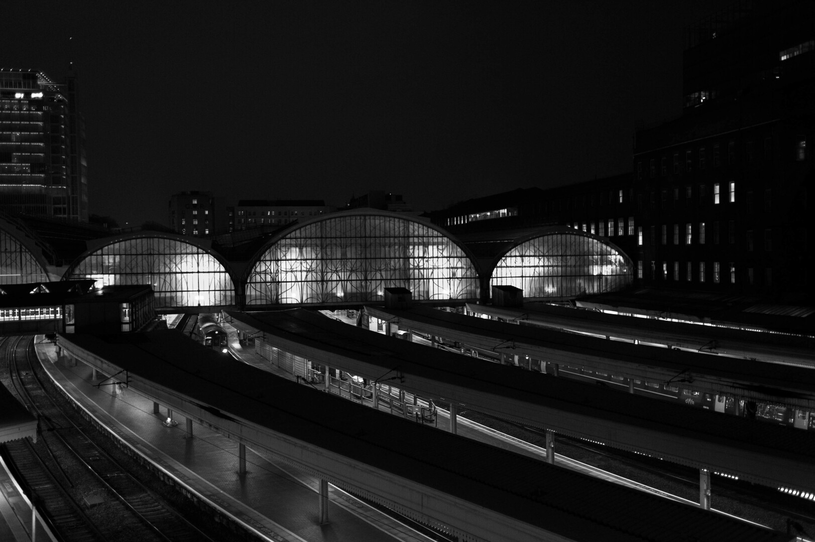 Night monochrome photo of several trainlines, covered with darkened canopies, under-lit, curving into a large train shed, roofed with broad arches, one substantially larger than the others, all glass-fronted, glowing from within. Behind this rise the rectangle lights of the city.