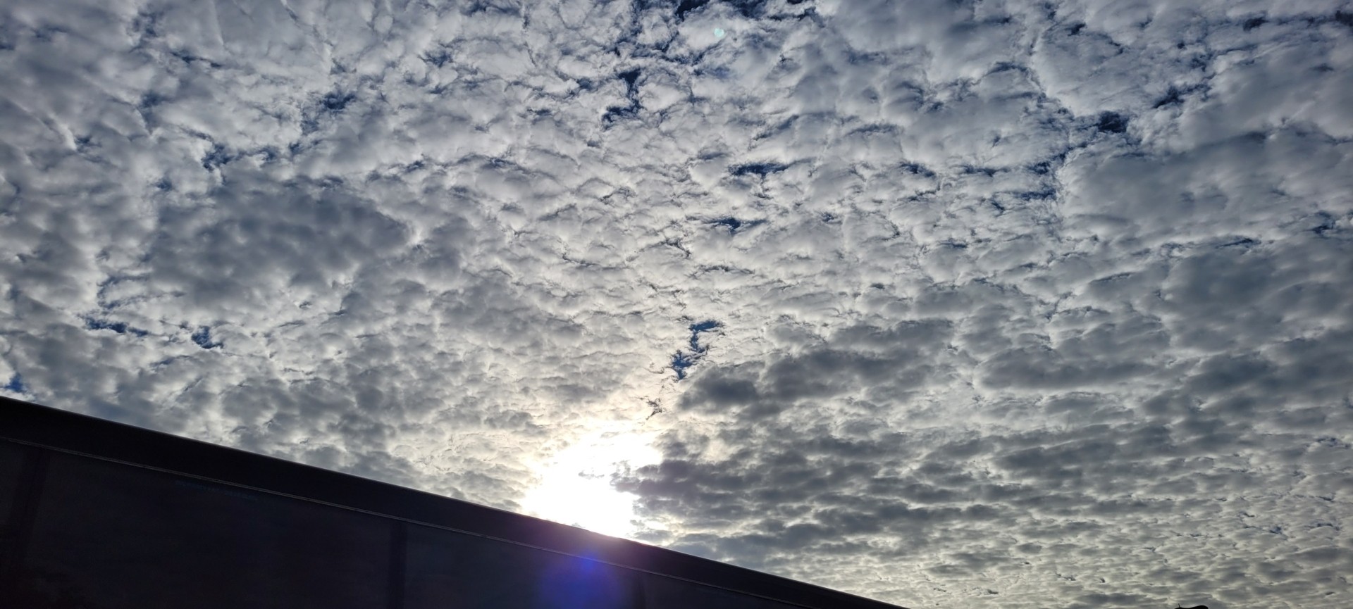 Across the dark angle of a roof, the sky is scudded with cloud cover brilliantly lit by a bright sun in a blue sky