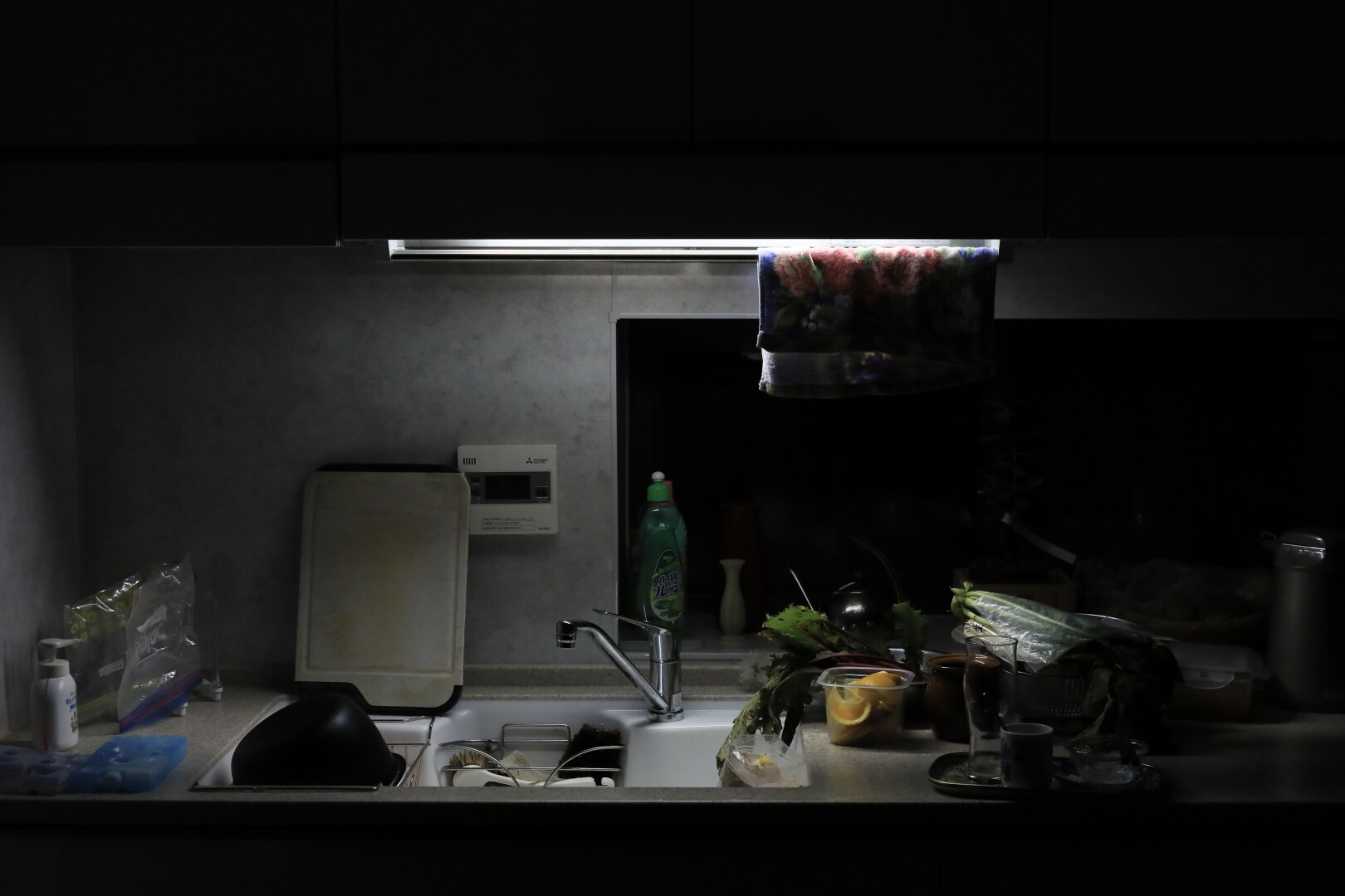 A cluttered kitchen sink is illuminated by a single cold LED lighting from above.