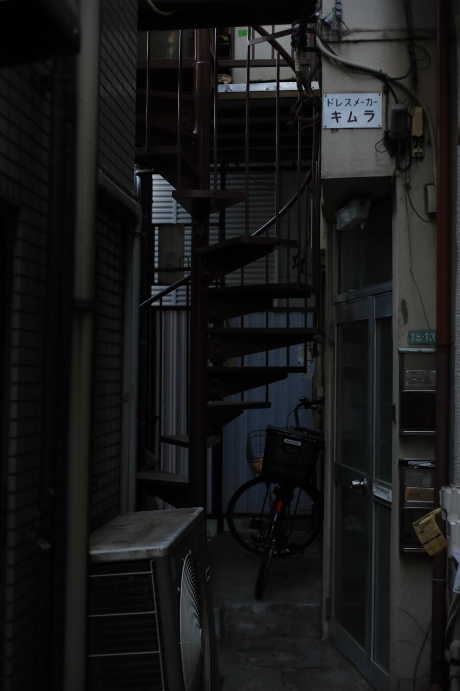 A dimly lit corridor between buildings.
A spiral staircase stretches upward, and a bicycle leans against a wall filled with mailboxes and signs.