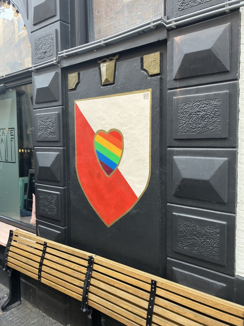 The arms of Utrecht, a shield with white top right and red bottom left, with superimposed a rainbow heart is painted on a wall. The painting is flanked by dark green stone cut in various geometric structures. A slatted bench is folded up against the wall.