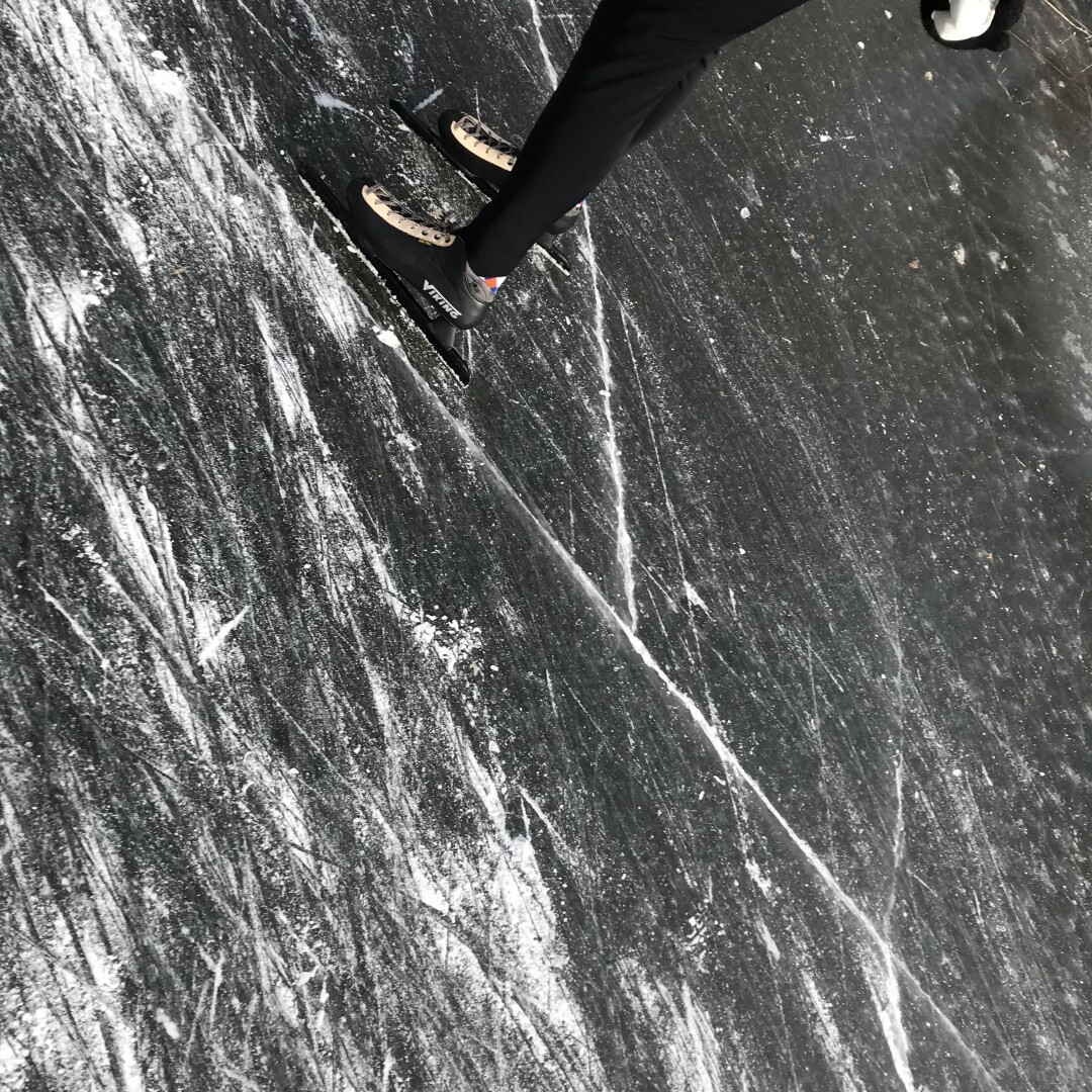 Tilted image of a dark frozen pond with skate marks and bright skate dust. In the top left the legs of a man in black leggings on speed skates are visible, appearing to skate out of the picture.