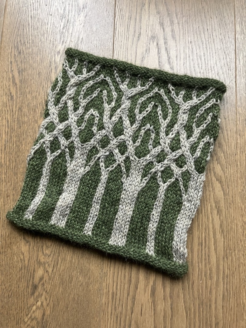 A knitted cowl in beige on a moss green background, looking like bare trees with intertwined branches