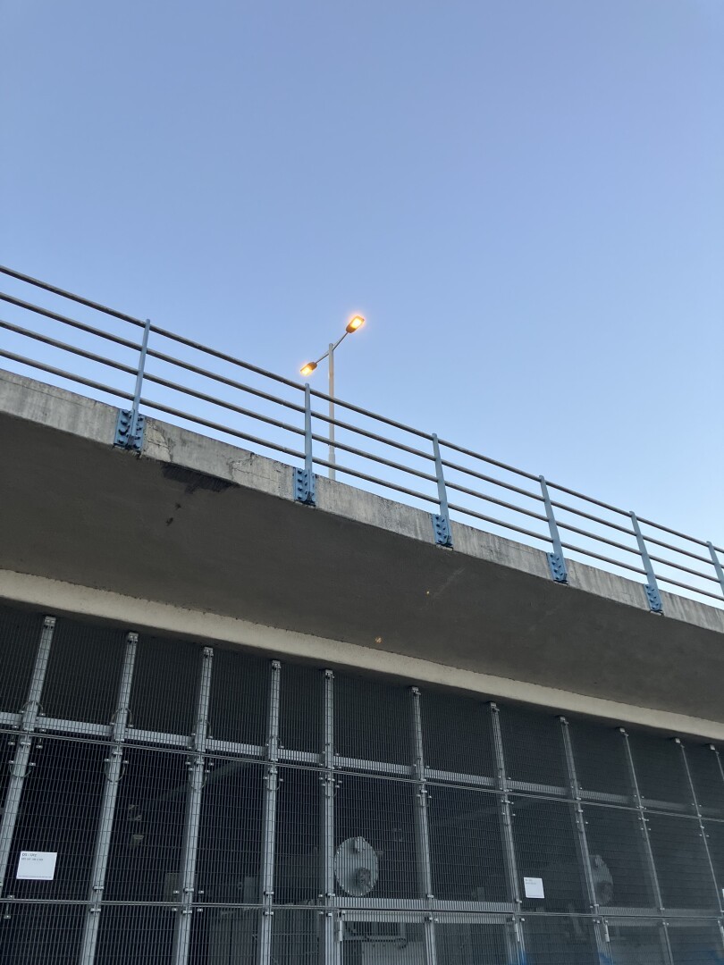 Street lights on an overpass, seen against a blue sky from ground level. The foreground is the fenced underside of the overpass.