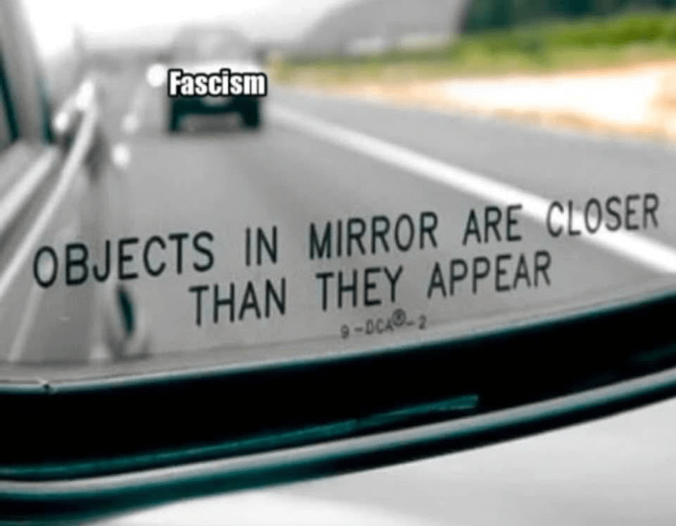 An image of a car mirror with a car in the background with the label "fascism&quot; and text below saying &quot;objects in mirror are closer than they appear&quot;