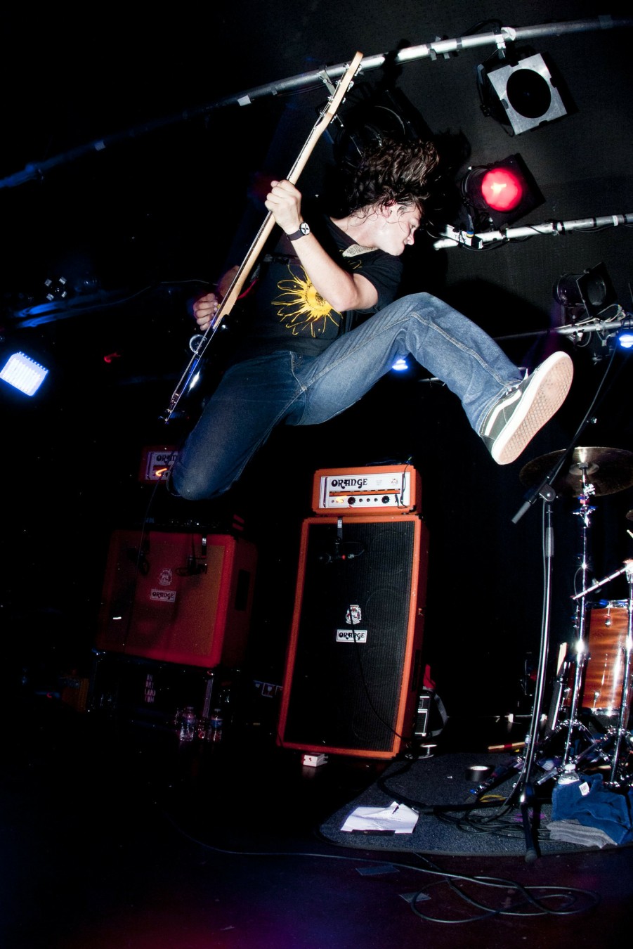 The singer/guitarist of Title Fight jumps high with his left leg outstretched to the side en his right leg is bend up. He is holding is guitar sideways above his head.
Behind him is his Orange Amp and Speaker.