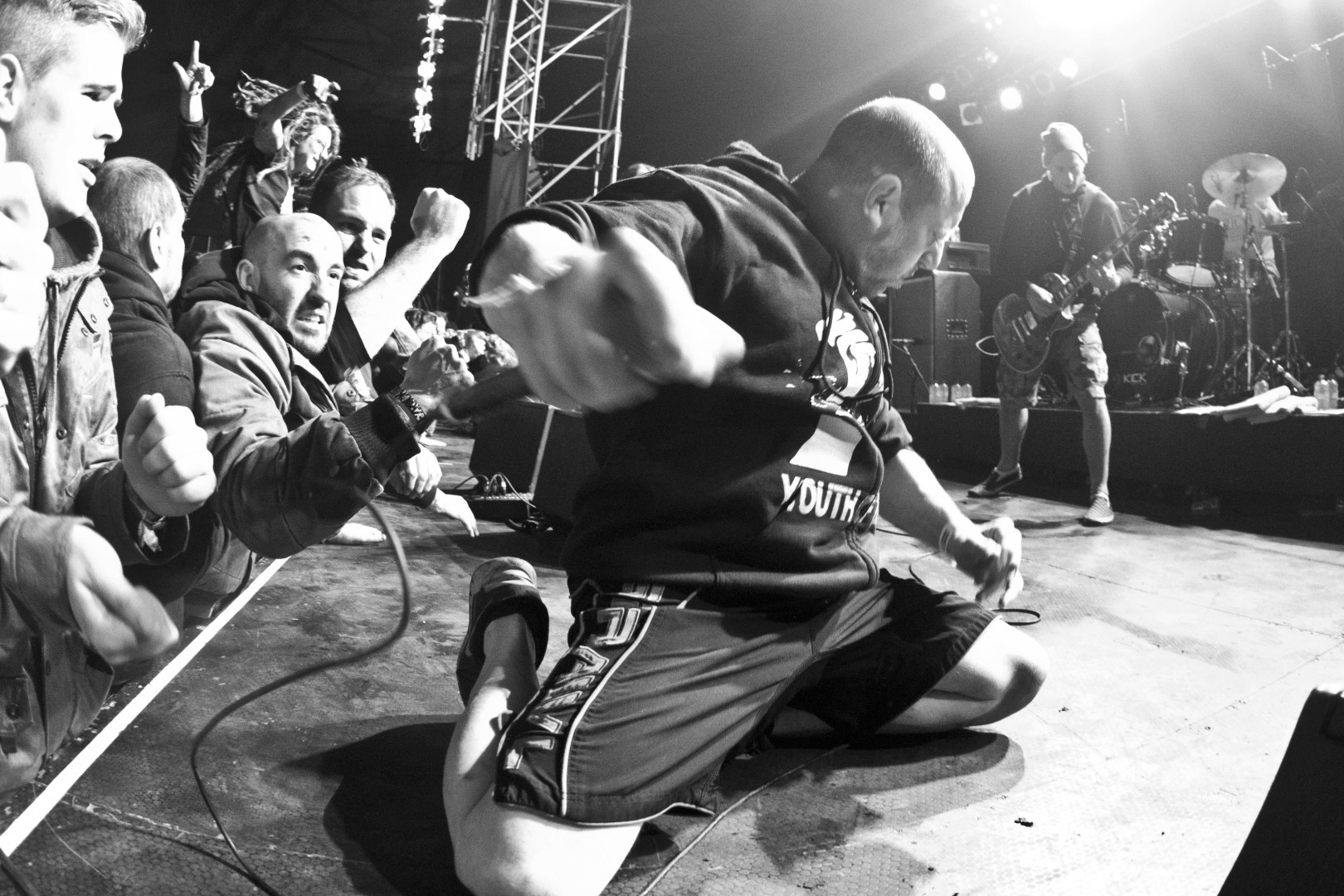 a photo of the singer of youth of today, Ray Cappo. he is on his knees on the front of the stage, fist with microphone almost in the camera. 
the crowd is push up against the stage on the left of the photo.