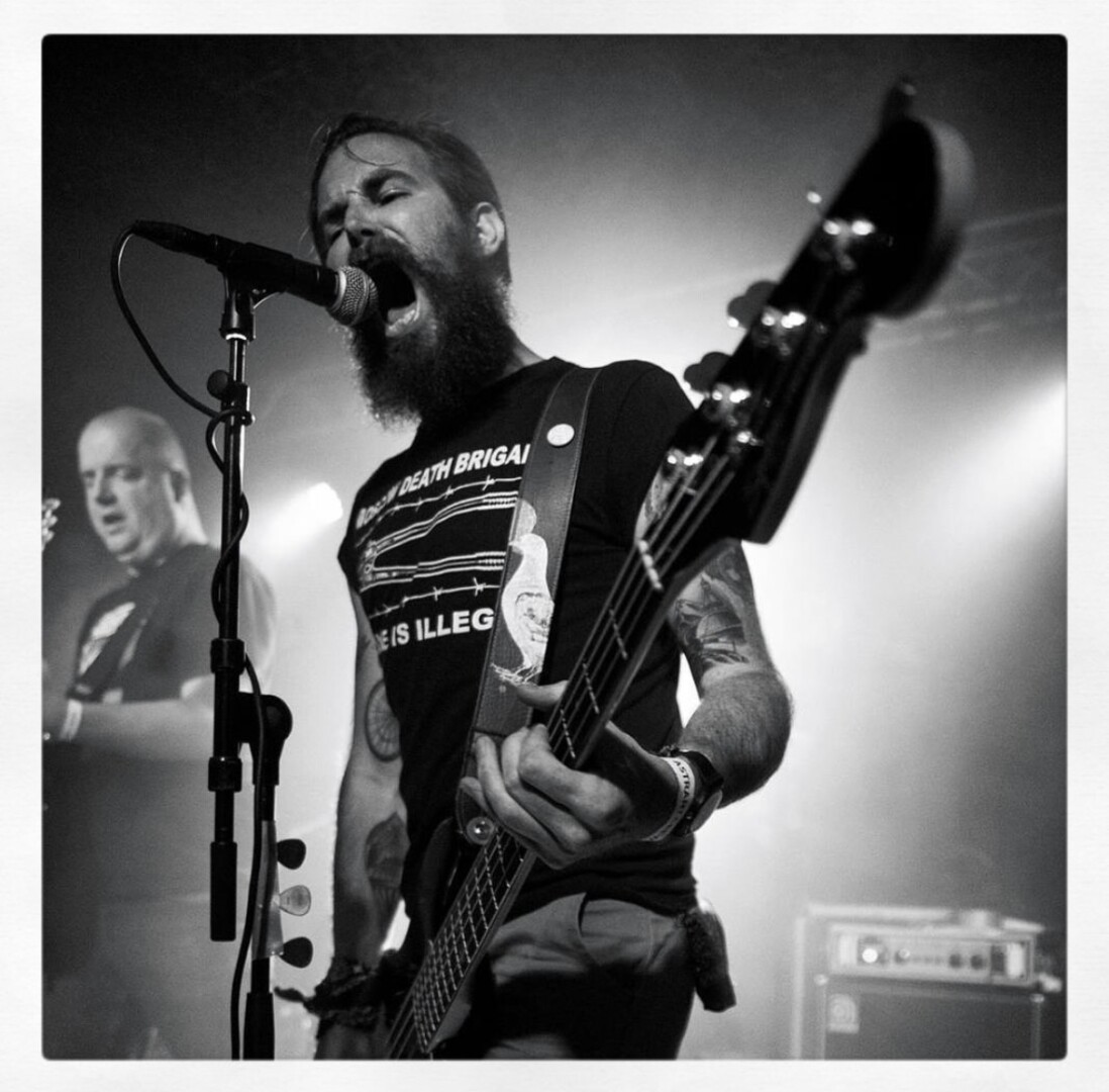 A black and white photo of a bass player with a beard and tattoos. 
He is yelling in the microphone while his bass fills a large part of the photo.