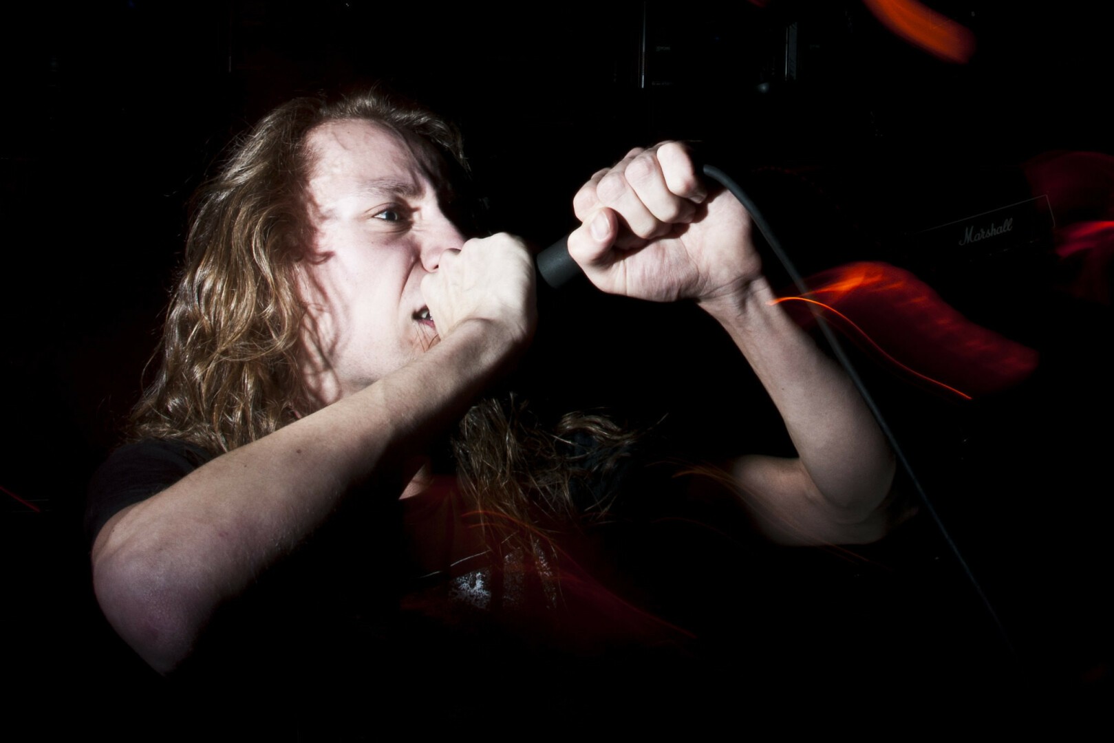 A hardcore vocalist (Naom Cohen) with long hair of this routine is hell holding his microphone with two hands while screaming into it.
Because of use of a sideways flash only his arms and face ar lighted while the rest of the photo is dark/almost black