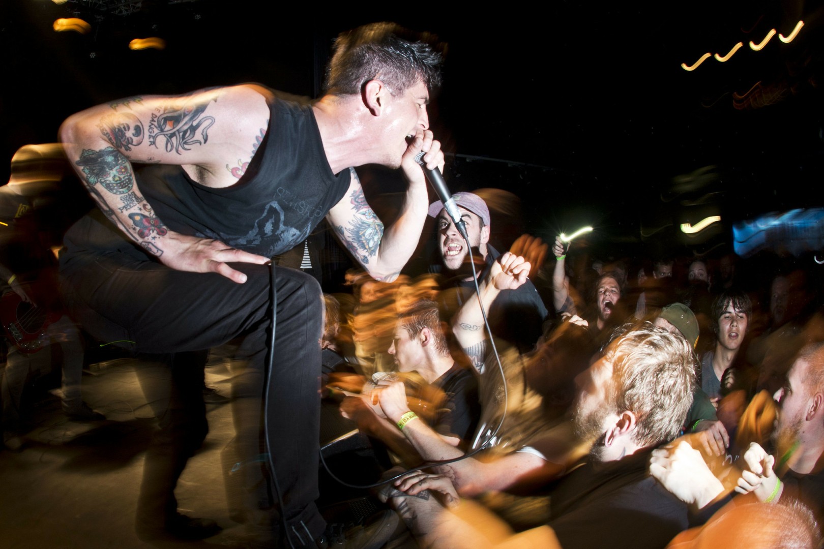 A photo of a singer - Jeffrey Eaton. He wears a sleeveless shirt, white arms with tattoo and is screaming in the microphone. His posture looks tense. He is screaming towards a front row which looks like a swirling mass of people.