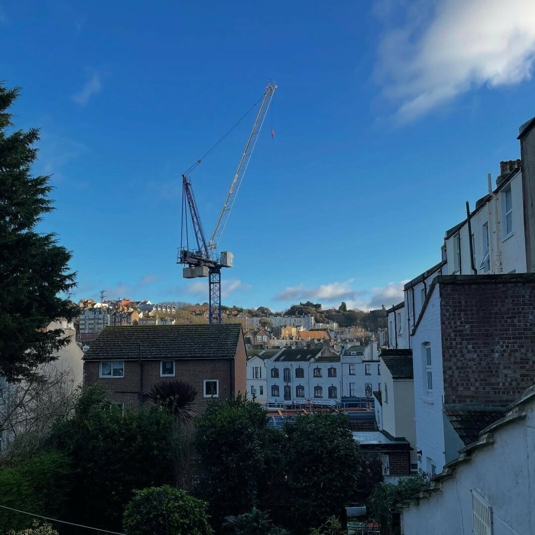 Tower crane visible against a blue sky between the edges of nearer buildings with a built up hill behind it.
