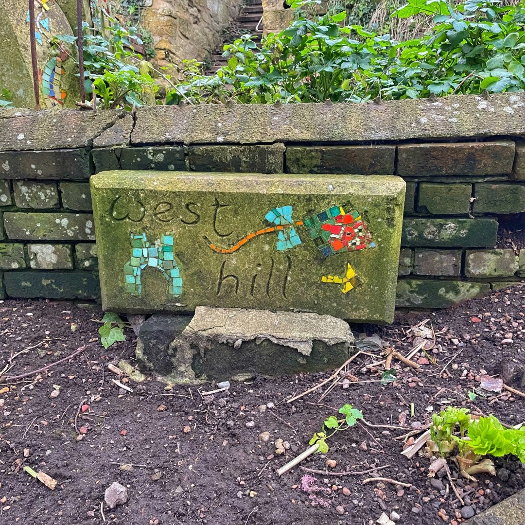 Upright landscape slab of concrete with the words West Hill drawn in it plus some elements of mosaic including an arrow pointing right