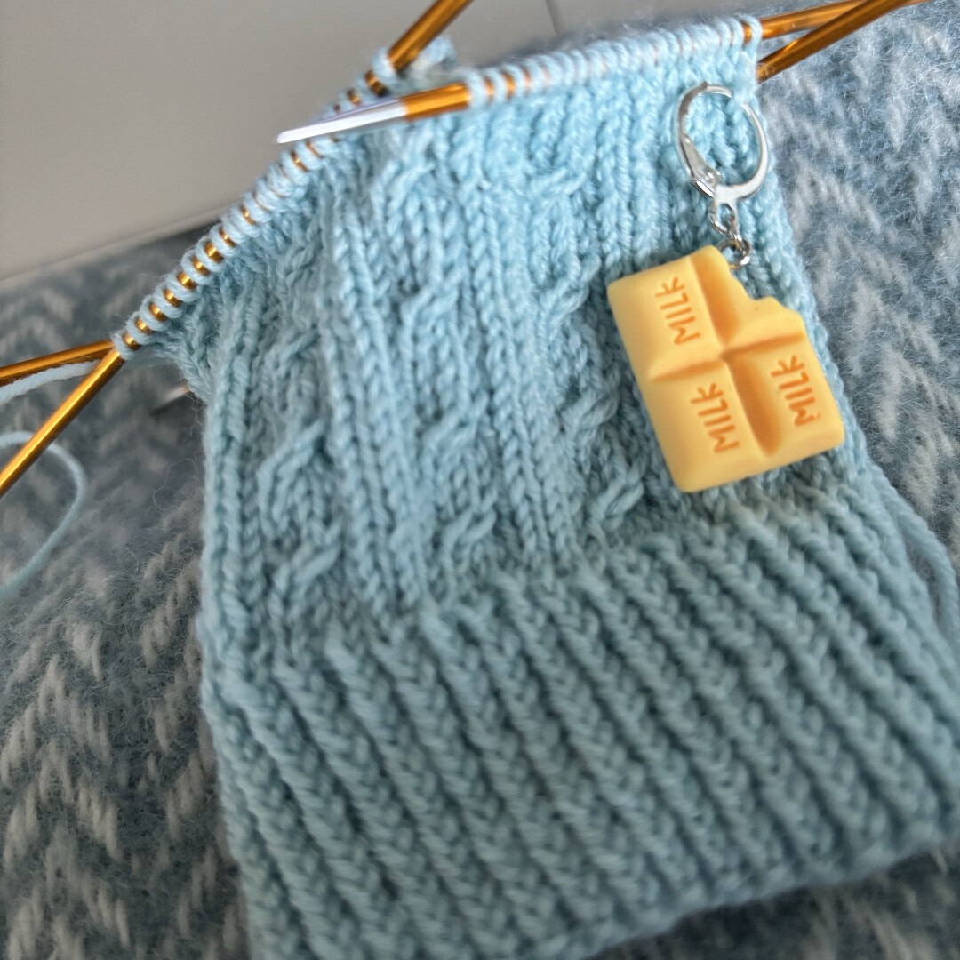 An ice blue sock in progress on orange dpns, with a small 3-stitch cable pattern. The cuff is in twisted rib. A progress keeper shaped like a slab of white chocolate is hanging a few rows below the needles.