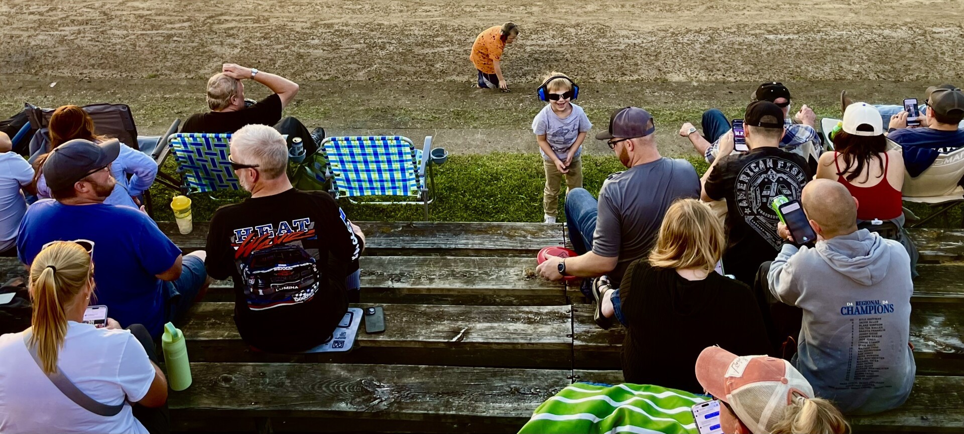 A group of people waiting for the races. A boy with ear protection, a girl in orange picking up stones, a blue/green lawn chair, adults talking to each other. The sun is low.