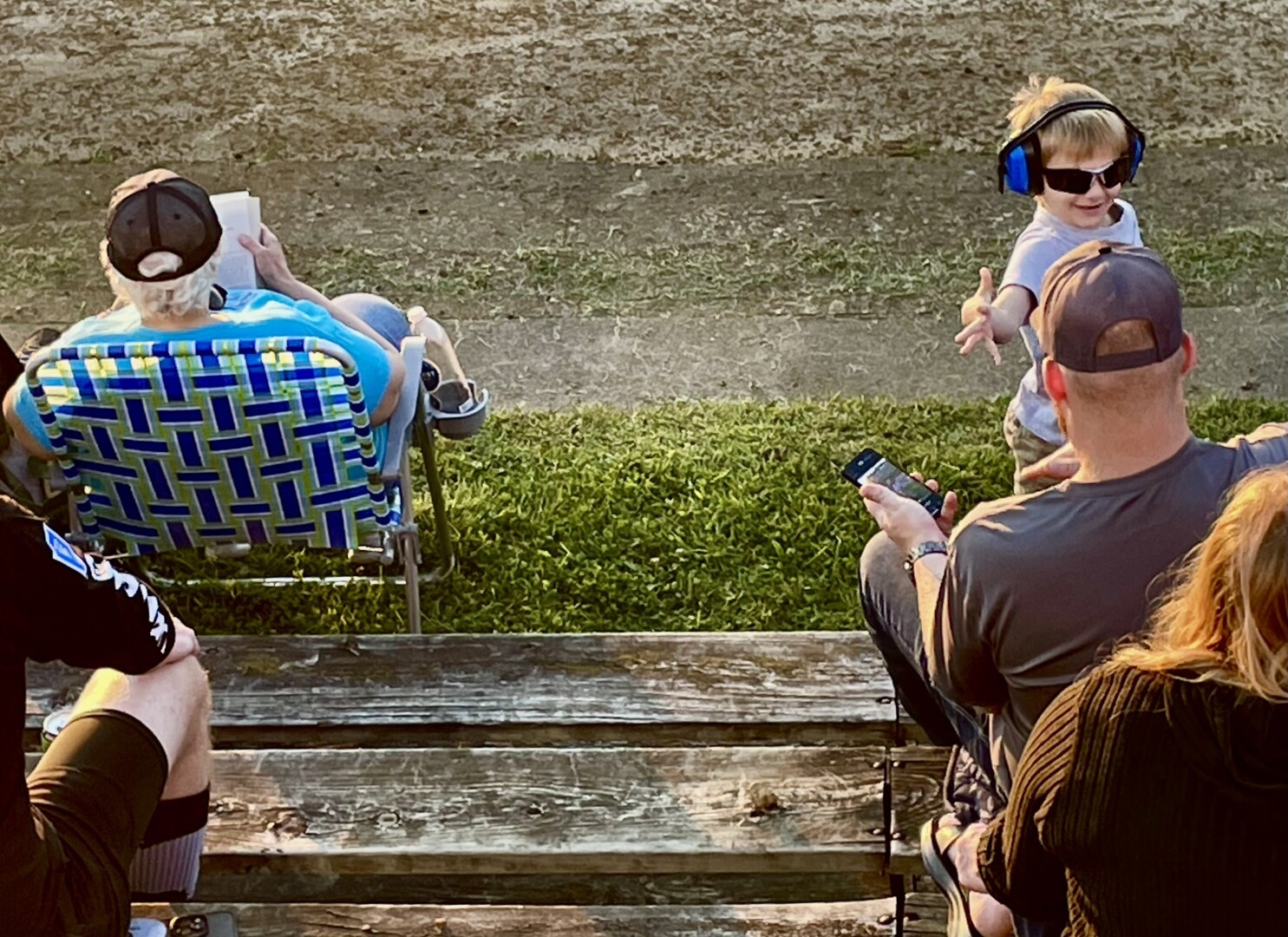 The perspective is a bit closer, so fewer adults wait. The ear protection boy wears sunglasses and gives a hand slap to a waiting adult. The races will start soon, the sun will set.