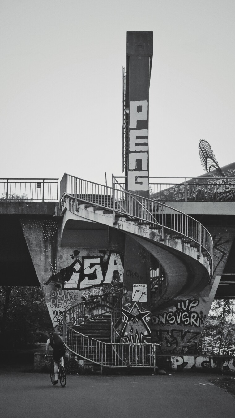 A grayscale, vertical shot captures a spiral staircase ascending from the lower left to the upper right, situated beneath a concrete bridge. The bridge is heavily covered in graffiti, the most prominent of which is a PENG tag. To the left of the staircase, a person on a bicycle rides away from the viewer. In the background, a metal railing and the underside of the bridge are visible against a light sky. A grayscale, vertical shot captures a spiral staircase ascending from the lower left to the upper right, situated beneath a concrete bridge. The bridge is heavily covered in graffiti, the most prominent of which is a PENG tag. To the left of the staircase, a person on a bicycle rides away from the viewer. In the background, a metal railing and the underside of the bridge are visible against a light sky.
