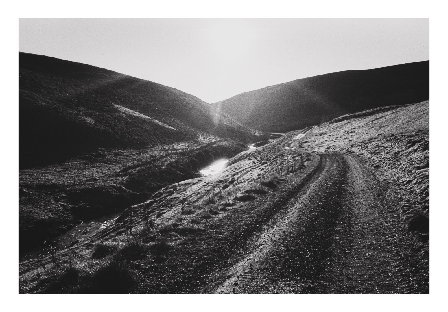 A black and white photograph of an upland scene. A track runs alongside a stream with hills descending on each side. On the horizon the sun sends distorted beams of light across the scene.