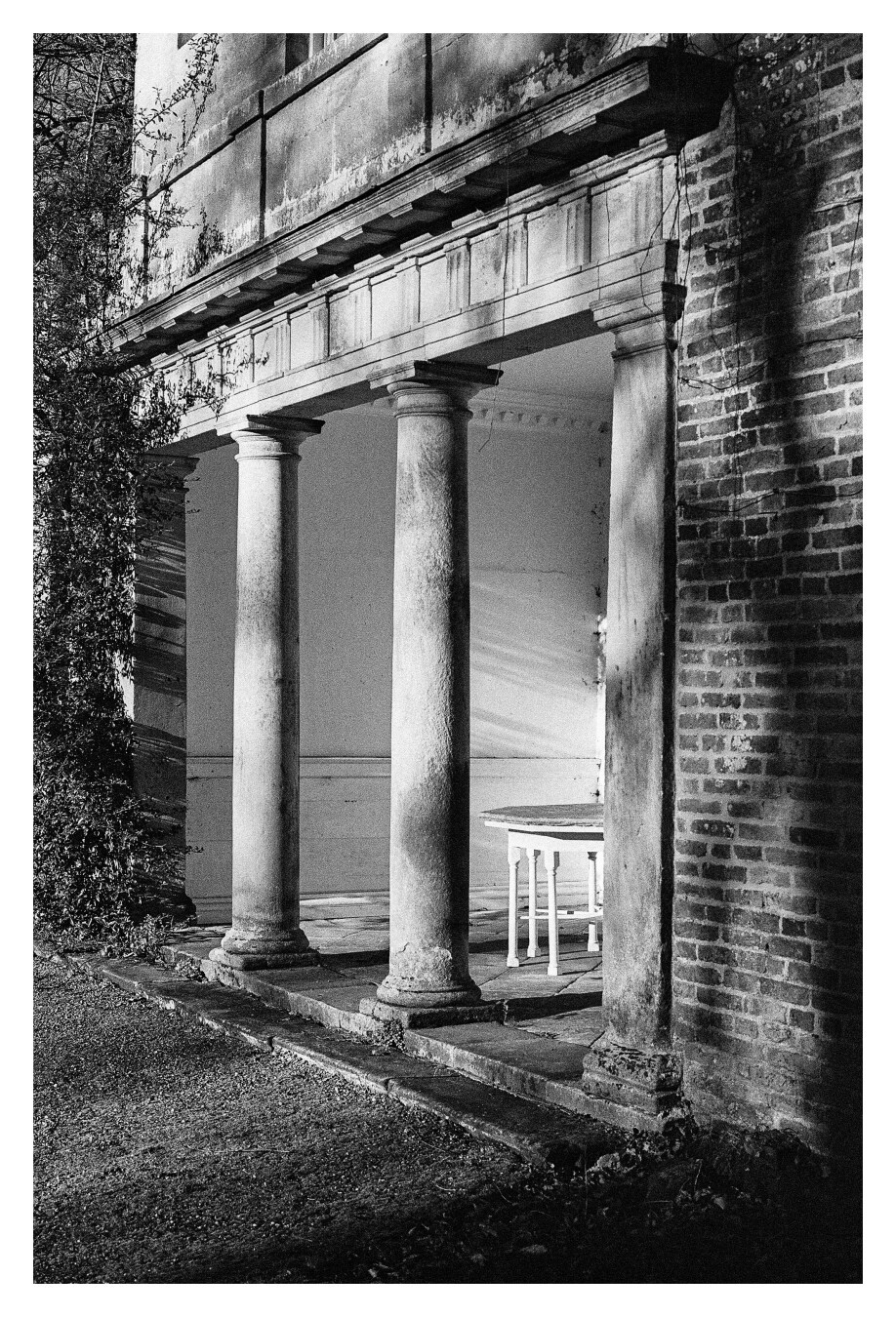 A black and white photograph of a small classical portico with three columns and a small picnic table in the recess.