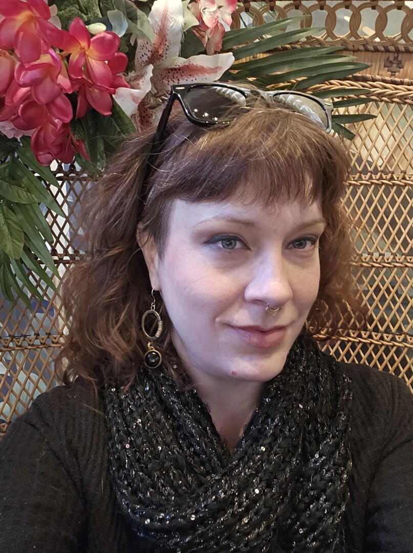 It me, a Caucasian lady in her 40s, dressed in black with a black knit scarf around my neck. I'm sitting in a high-backed ornate bamboo and wicker? chair adorned with faux flowers. There's a black pair of sunglasses in my hair, silver and brass circular asymmetrical geometric earrings dangling from my ears, and a gold septum ring in my nose. My hair is shoulder length, leans auburn-toned brown, and I have thick brow-length face-framing fringe.