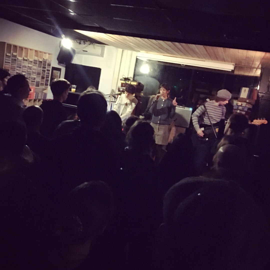 Foto van achter uit de zaal, over de hoofden het publiek heen. Op een klein podium staan drie mensen die muziek maken. Locatie is klein platenzaakje.￼