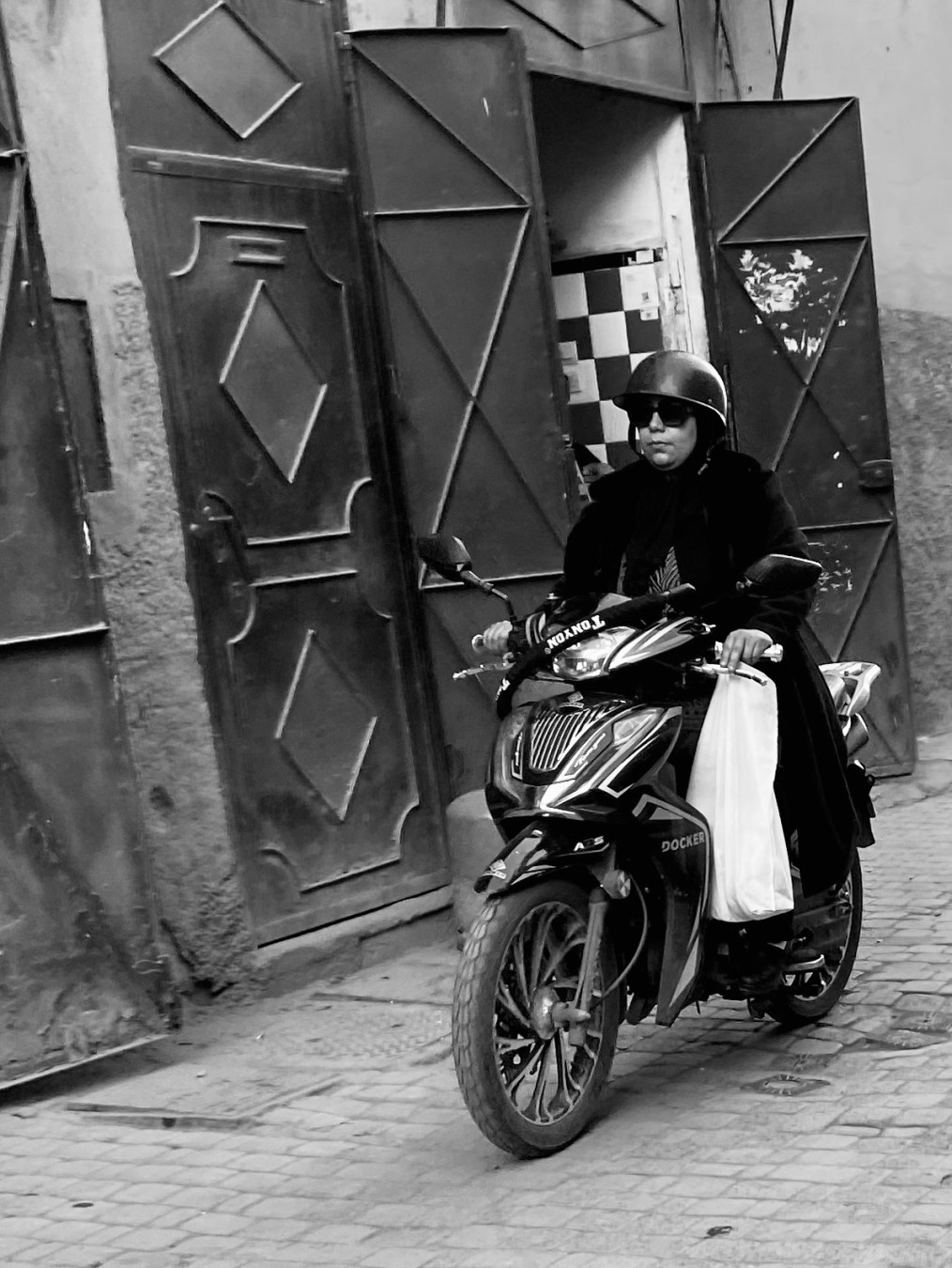 Woman in black helmet and sunglasses, carrying white shopping bag on scooter. (b/w)