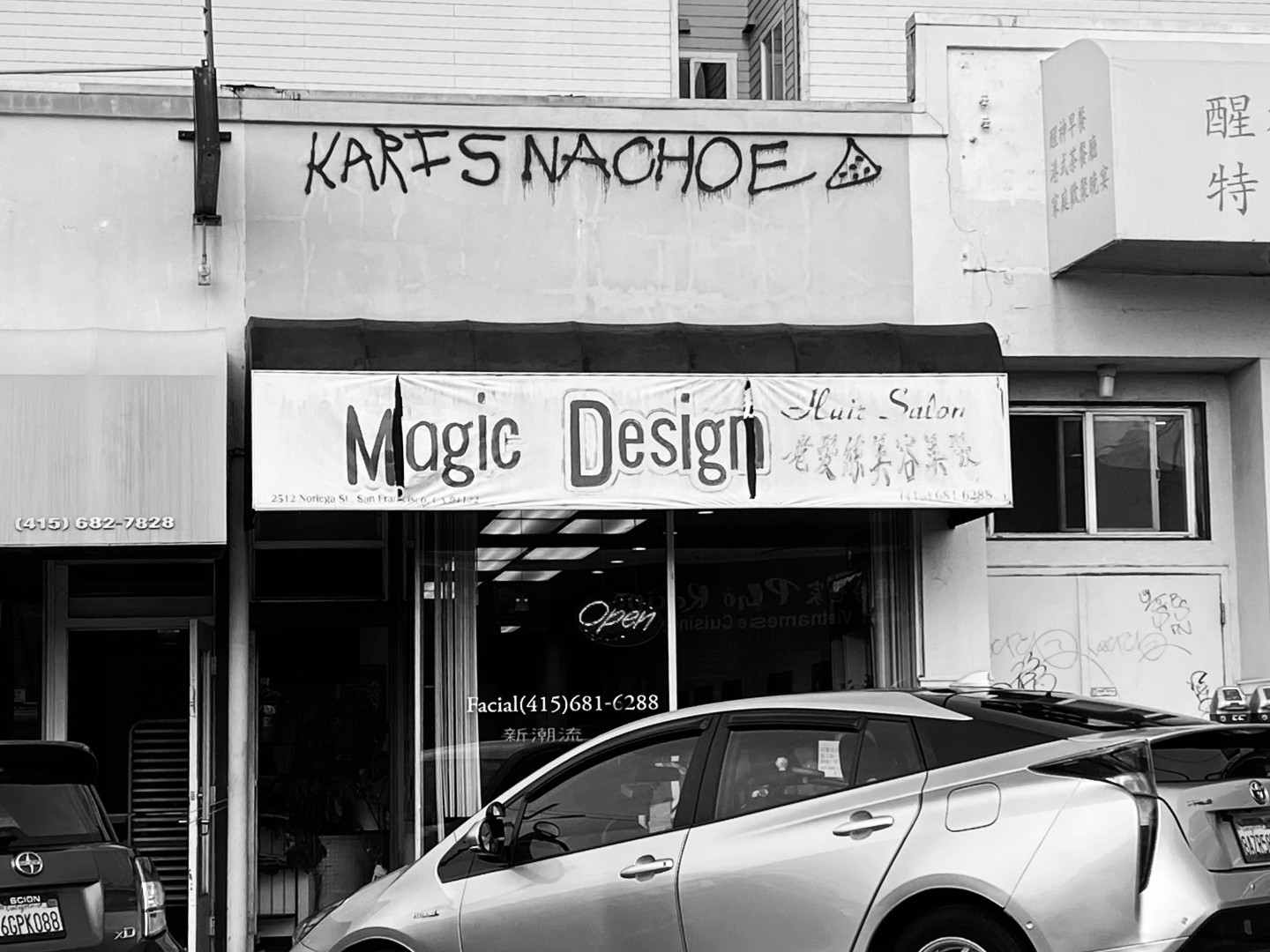 B/W street photo of salon with awning, “Magic Design Hair Salon,” with vertical rips between M and a, and in the final n. Above the awning on the building facade is spray painted fairly amateurishly, KARIS NACHOE, with a triangle at the end that’s apparently a corn chip but more resembles a pizza.