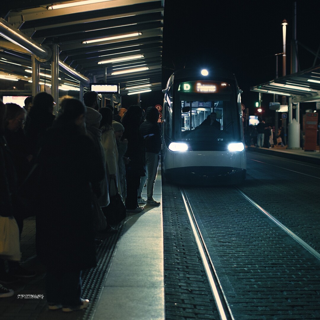 De nuit, un tram arrive en station. La photo est prise telle que le quai et sa toiture dessinent une ligne droite verticale au milieu de la photo. Le côté gauche de la photo est rempli d'une foule de dos, sombre et anonyme, tournée en direction du tram; tandis que le côté droit symbolise un grand espace vide avec les rails et le tram également sans passager.