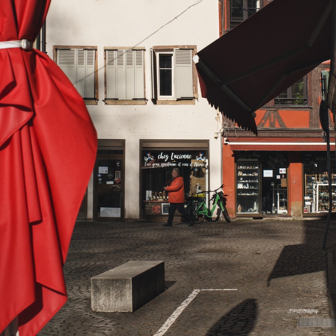 Une scène de rue, axée sur le rouge. Sur le côté gauche un parasol rouge replié. Au fond des immeubles dont celui de droite est à colombages et rouge. Au centre, un homme à l'anorak rouge traverse la scène.