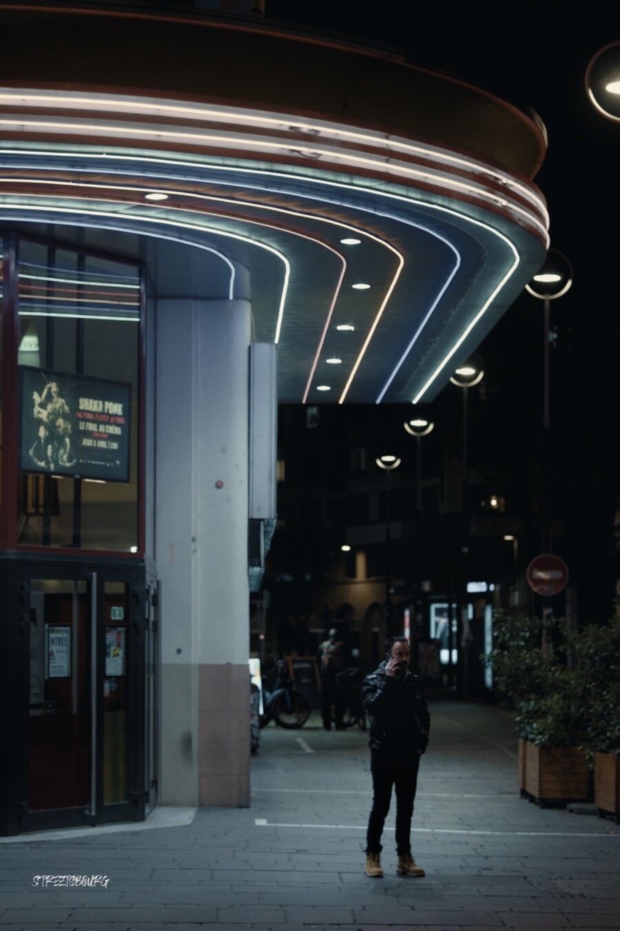 Une plan large de nuit à proximité de l'entrée d'un cinéma. Un homme se tient debout au téléphone. Au dessus de lui des néons colorés dessinent des arcs de cercle.