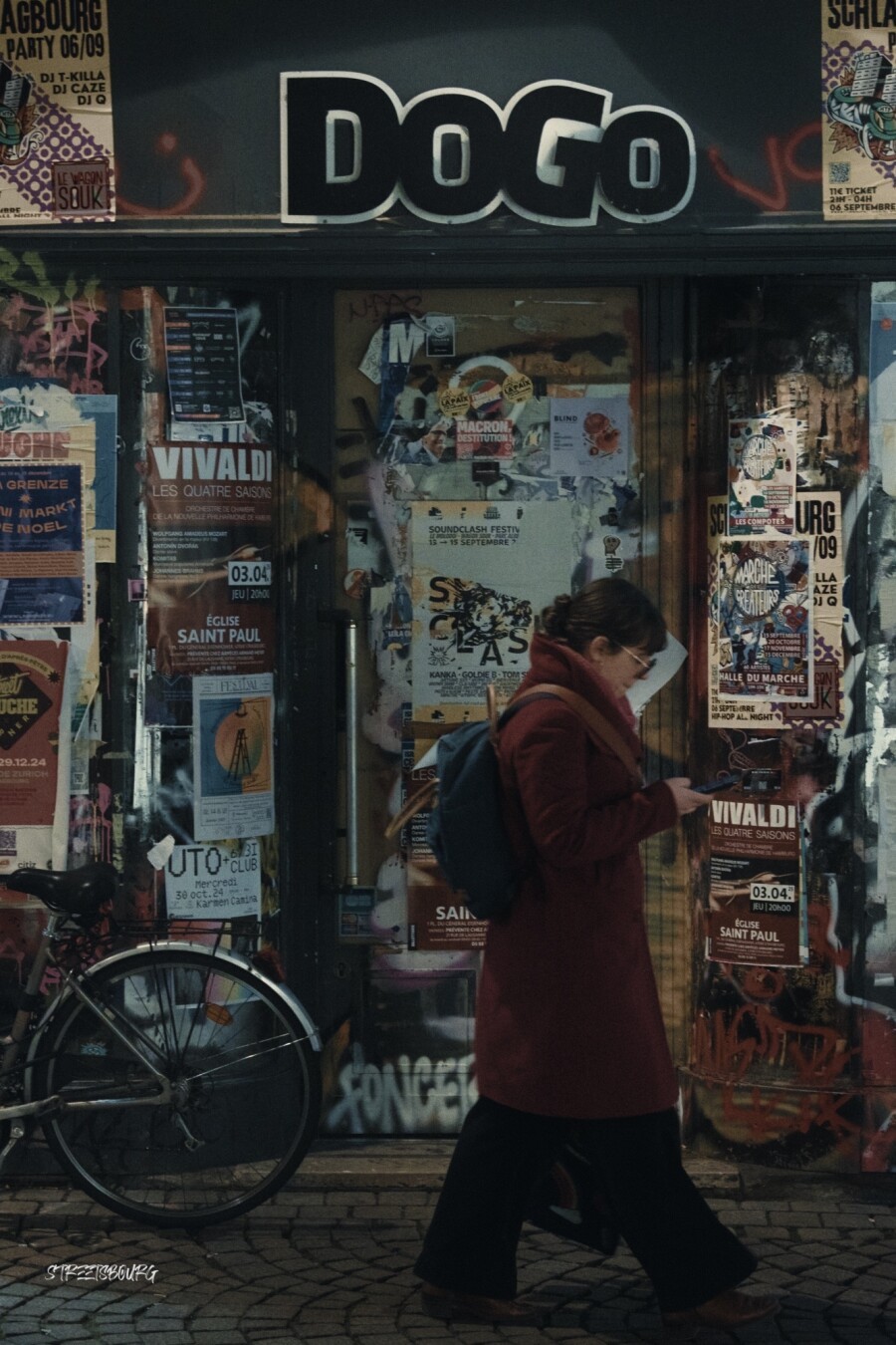 Une femme qui regarde son téléphone, passe devant une devanture «Dogo» qui semble abandonnée, car remplie d'affiches diverses collées. Sur la gauche est posé un vélo dont on ne distingue que la roue arrière