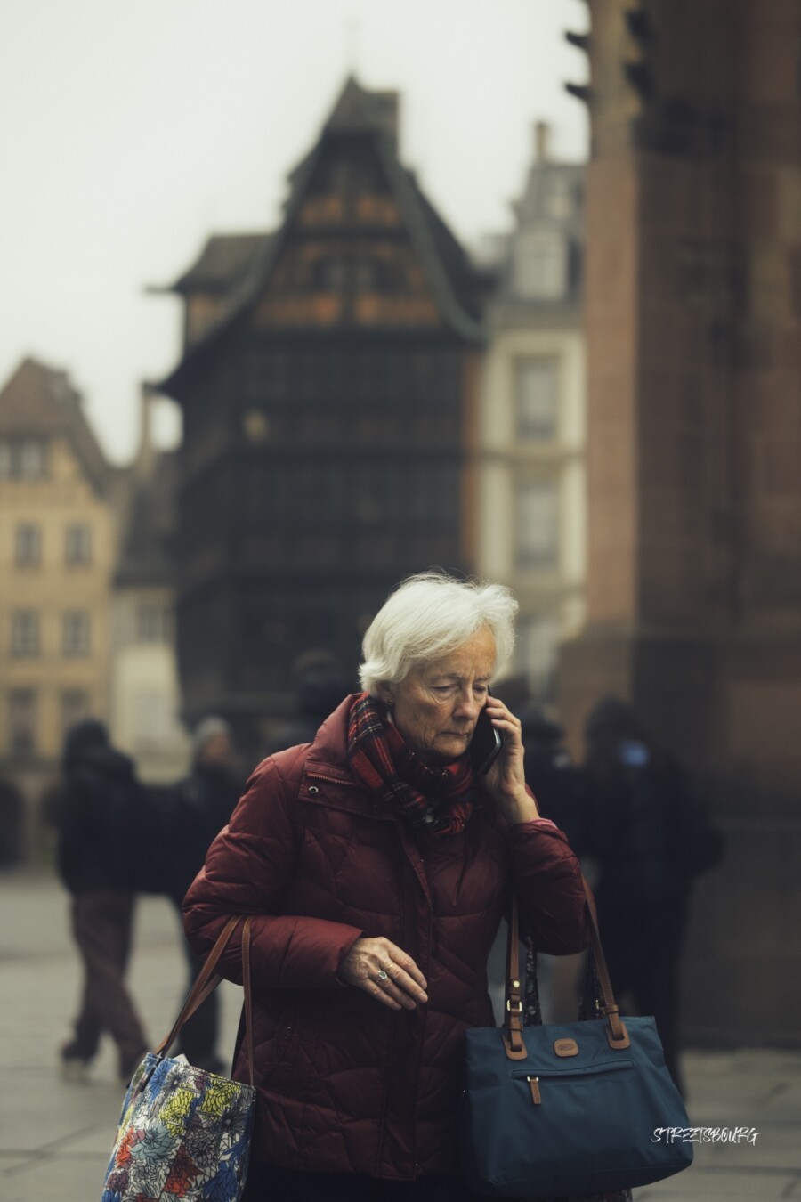 An old lady passing by, white hairs, closed face. She holds three handbags, while phoning. Behind in the distance is the Kammerzell house, the oldest house of Strasbourg