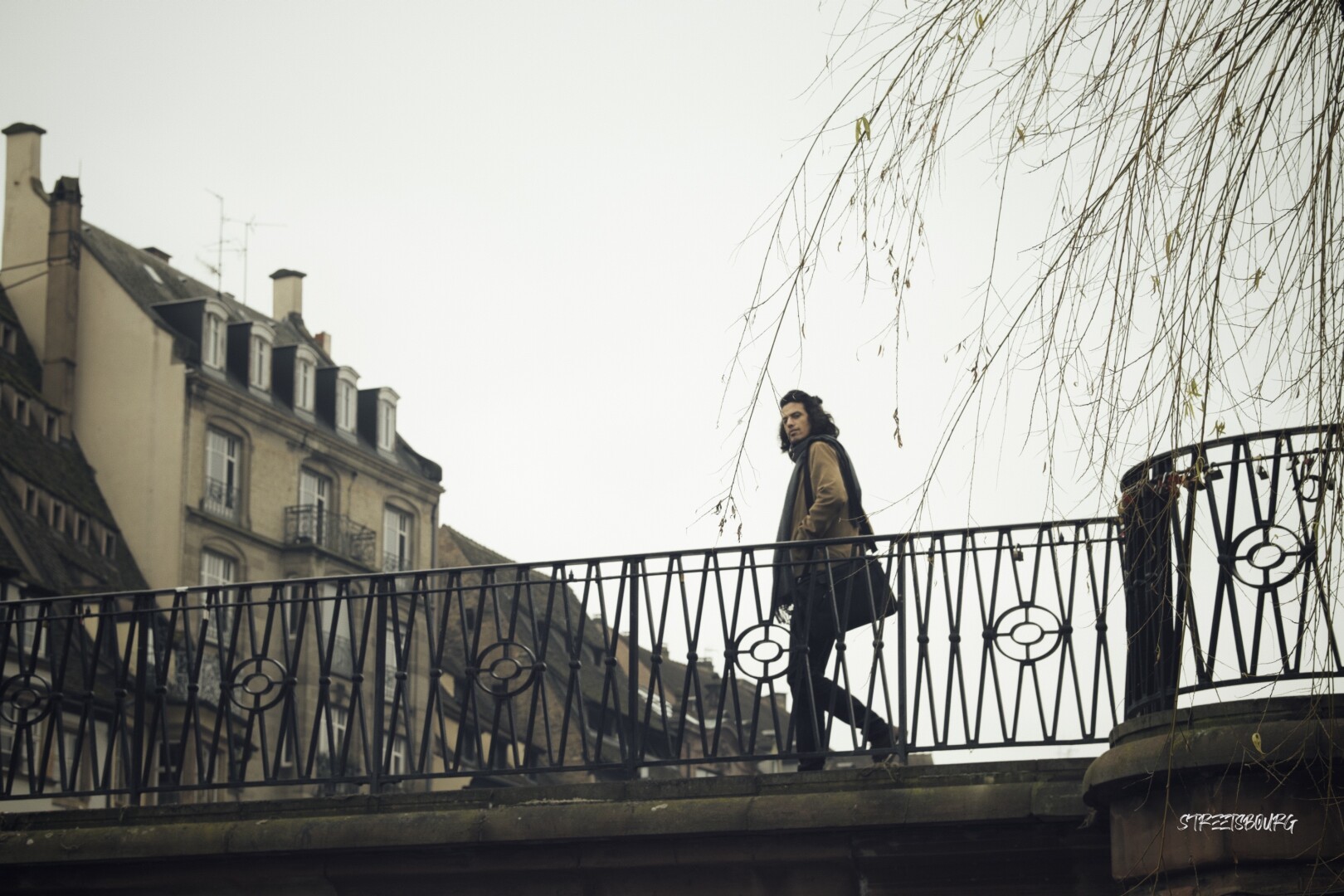 A man is passing by on a bridge, looking at the photograph. The photo is taken upward from the side of the river