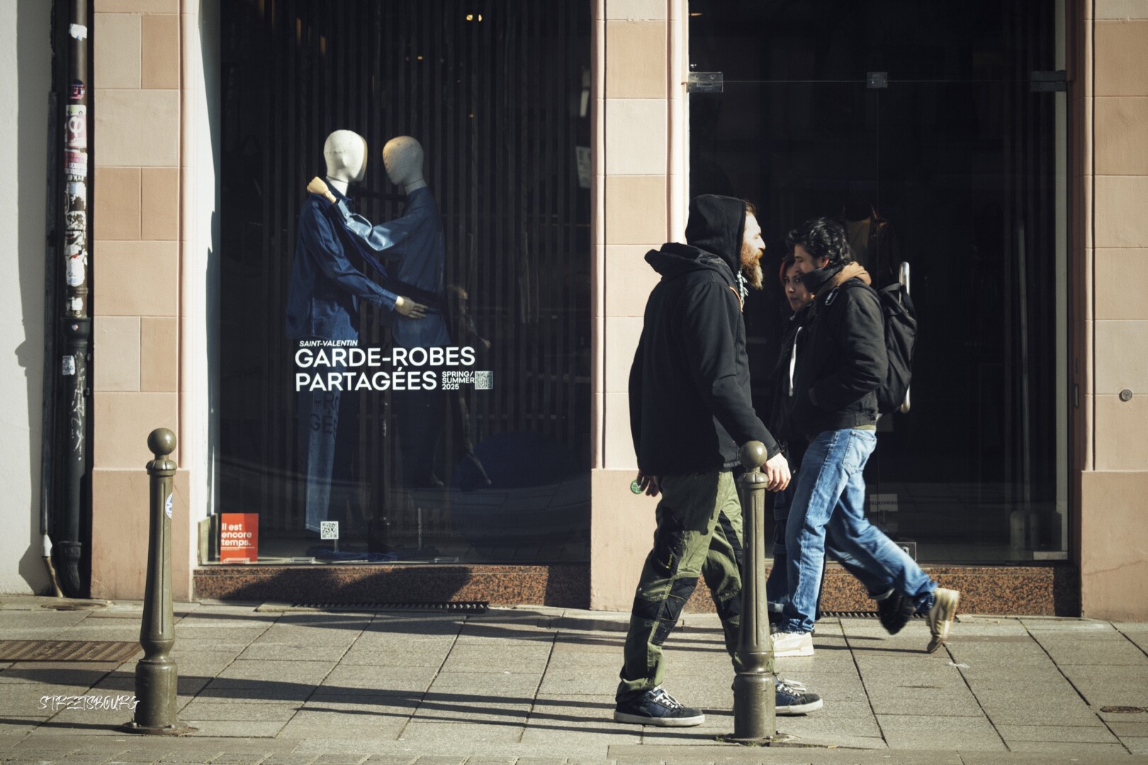 In front of a shop window displaying two mannequins standing as if to slow dance, a couple walks in one direction while a man walks in the other direction. The distance between the man and the couple is the same as between the mannequins. The woman in the couple looks at the man who is walking in the other direction.