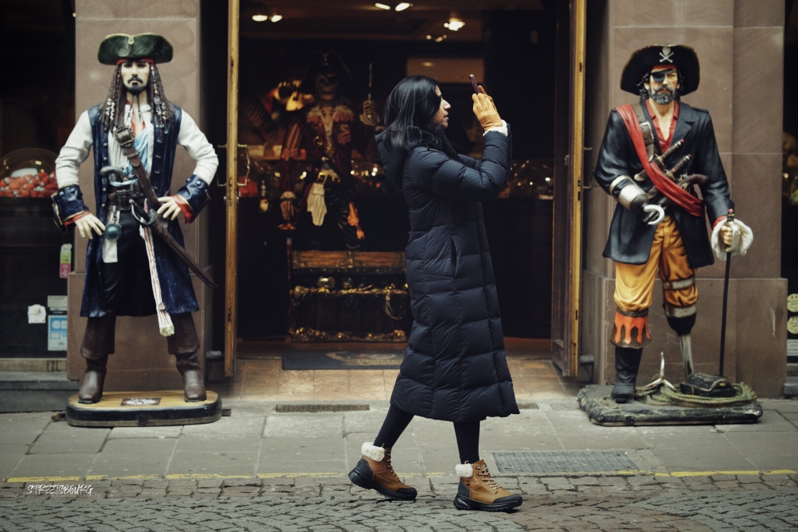 A woman in the center of the frame is facing right and pointing her phone at something for a picture. She stands between two pirates statues which are placed at the entrance of a candy shop