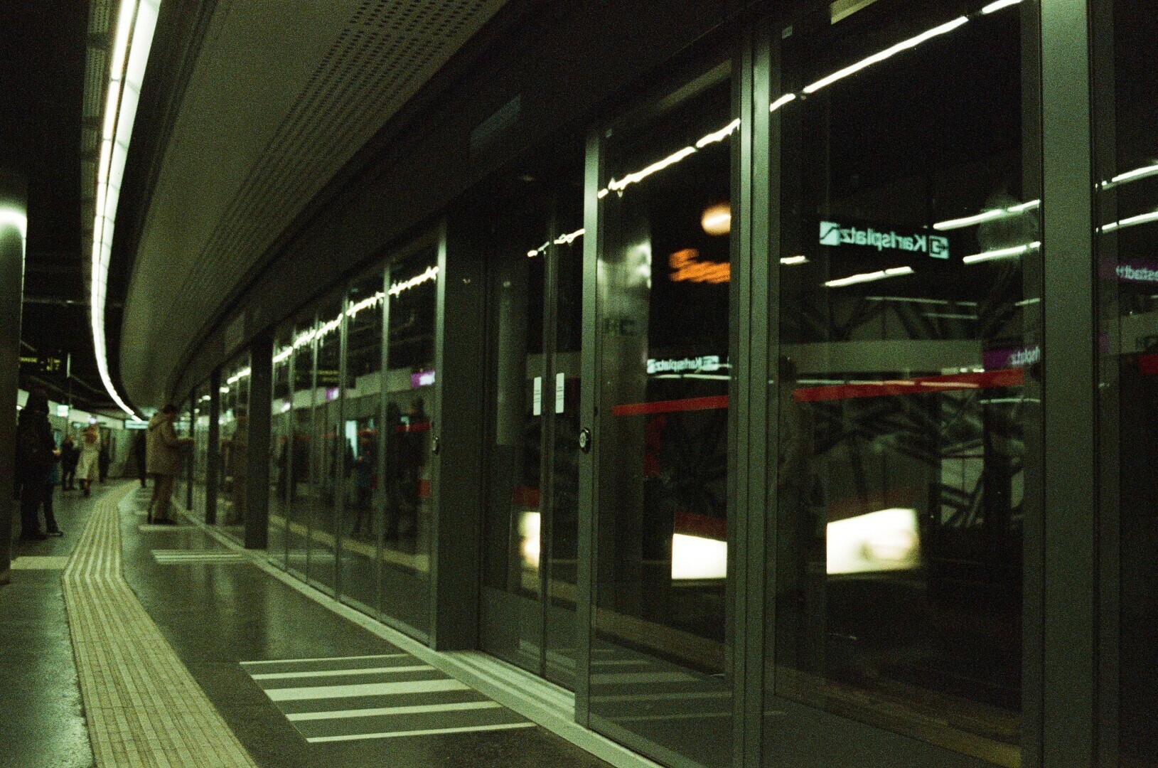 Photo of a curver underground metro station platform with glass screen dooors, bheind them train lights are seen. People are standing on the far side of the platform