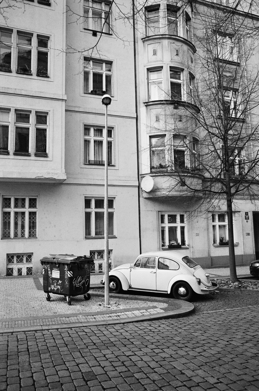 A monochrome photo of a street with sideways prewar building and a parked vw bug next to a full dumper