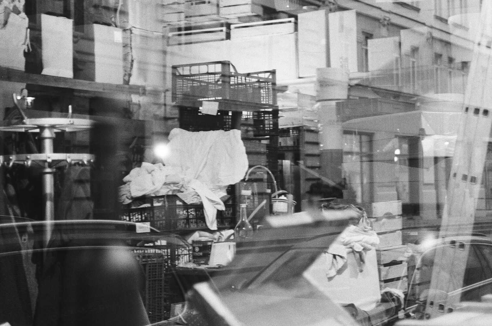 A monochrome photo of an interior of a closed shop with different boxes and unorganized stuff inside with slight reflections on the glass