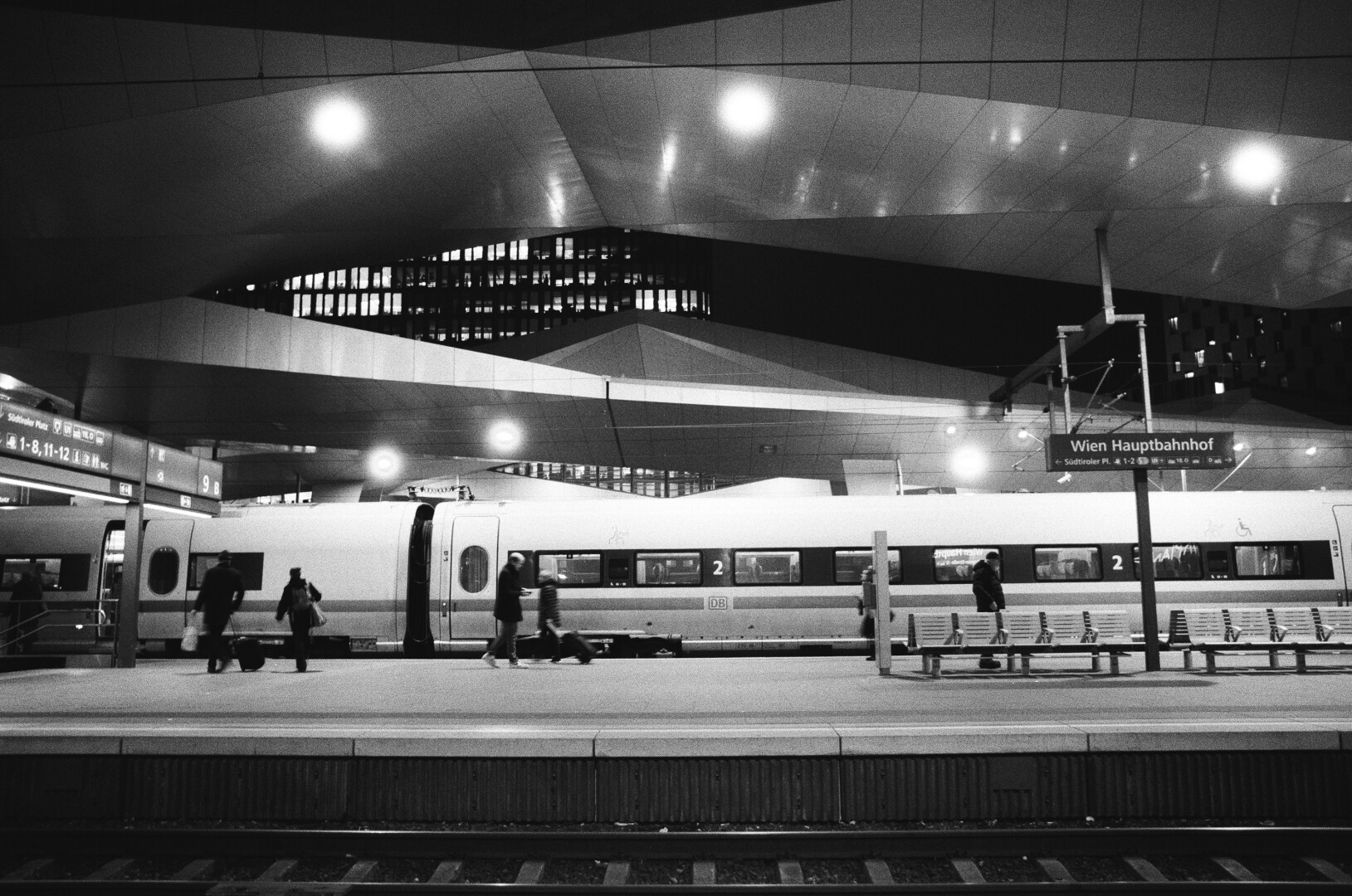 A monochrome photograph of a Wien HBF station platform with white DB train car with multiple people walking about.